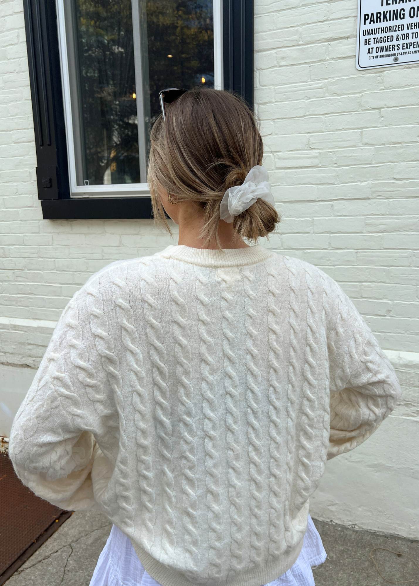Woman with blonde hair wearing the statement scrunchie in white and a cream coloured cable knit sweater in front of a white brick wall