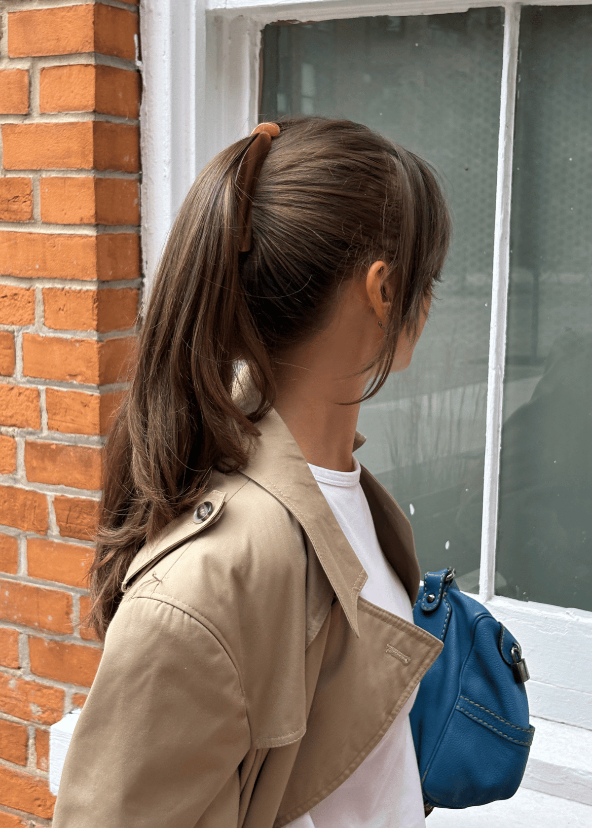 A woman with her hair in a high ponytail secured by a Banana Clip, wearing a beige trench coat over a white t-shirt and carrying a blue shoulder bag, standing next to a brick wall and a window.