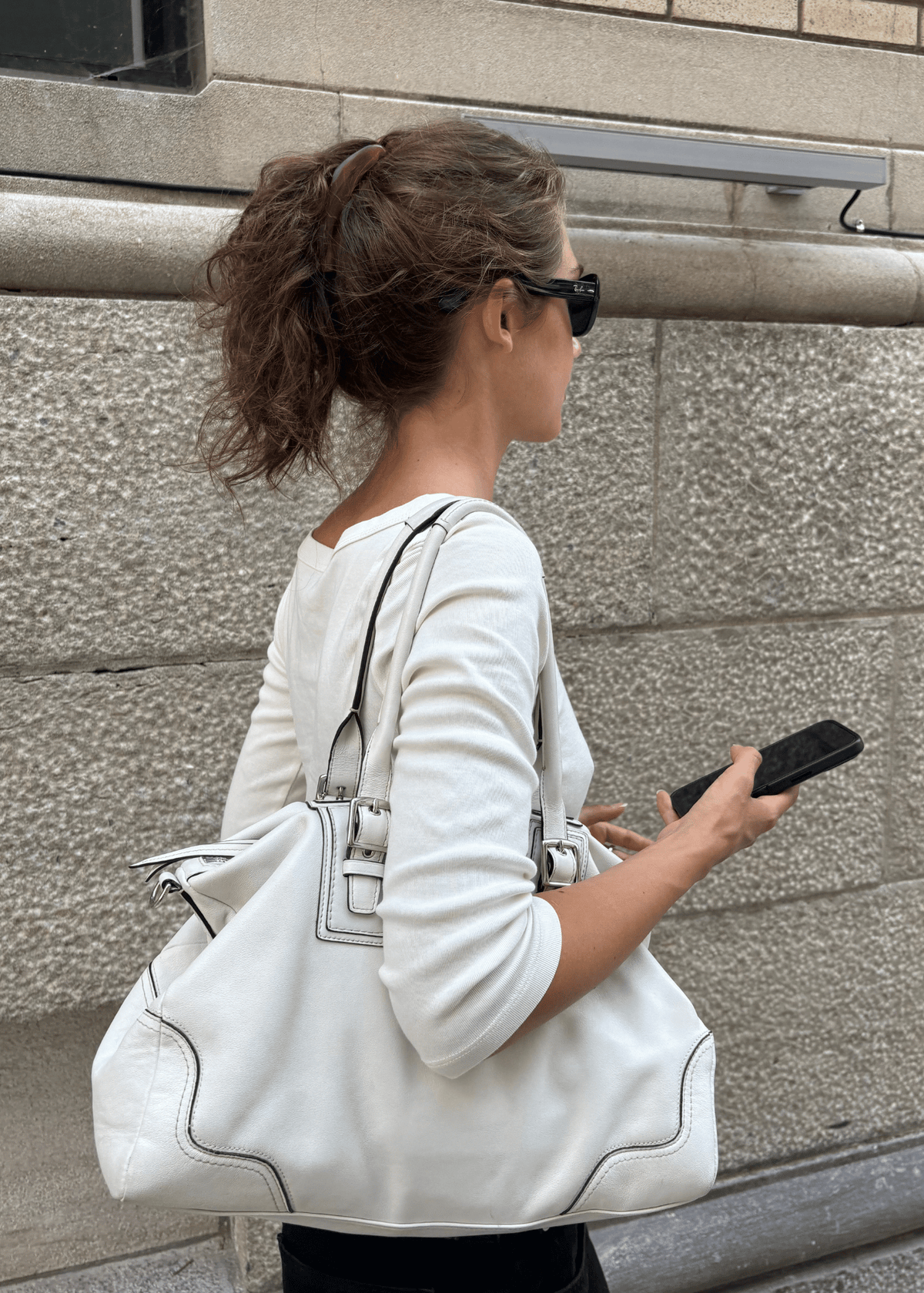A woman in a white long-sleeve top and black sunglasses carries a large white leather shoulder bag with black trim and stitching, standing against a gray stone wall. She wears her hair high on her head in a Banana Clip.