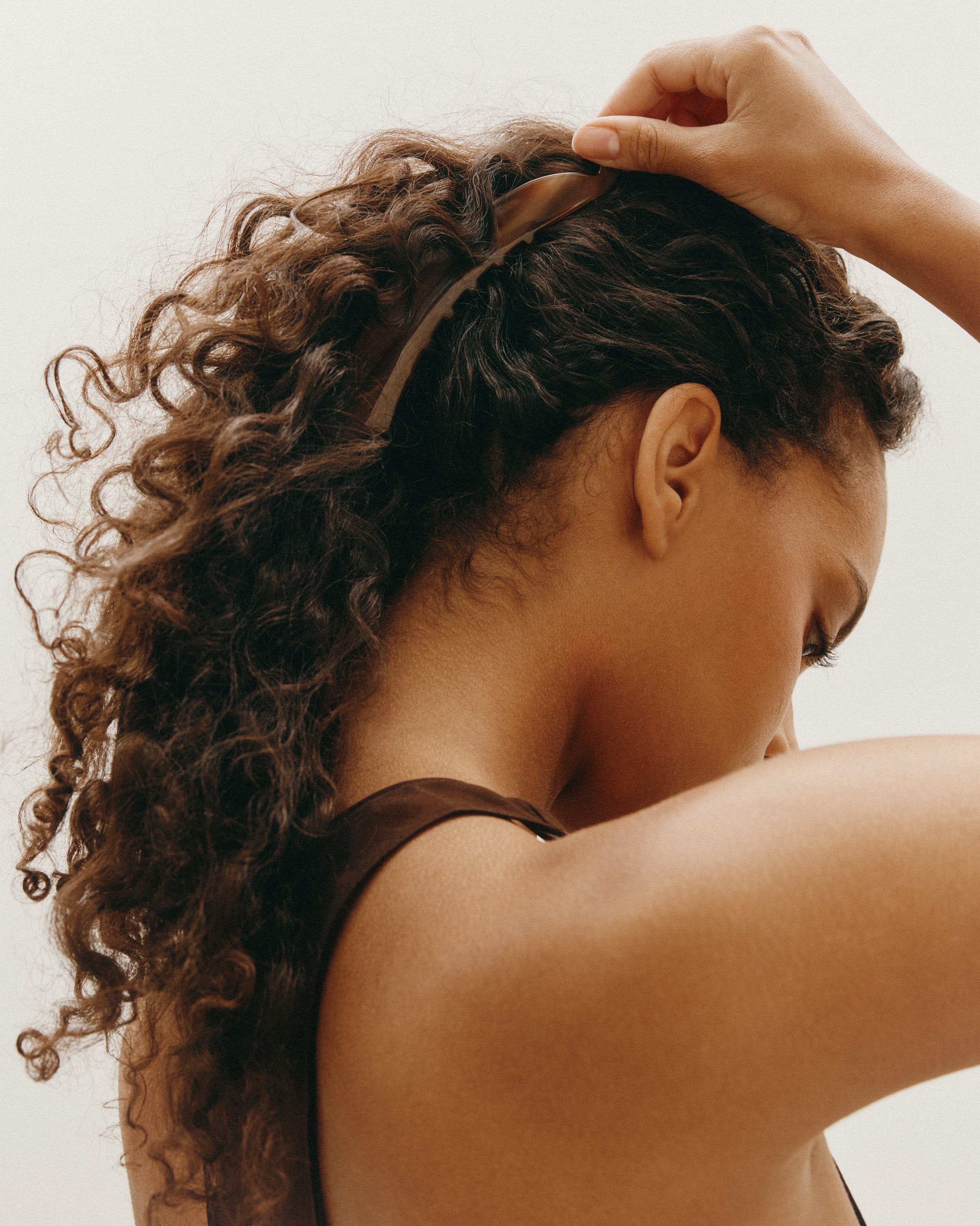 Woman adjusting a banana clip with curly brown  hair against a neutral background