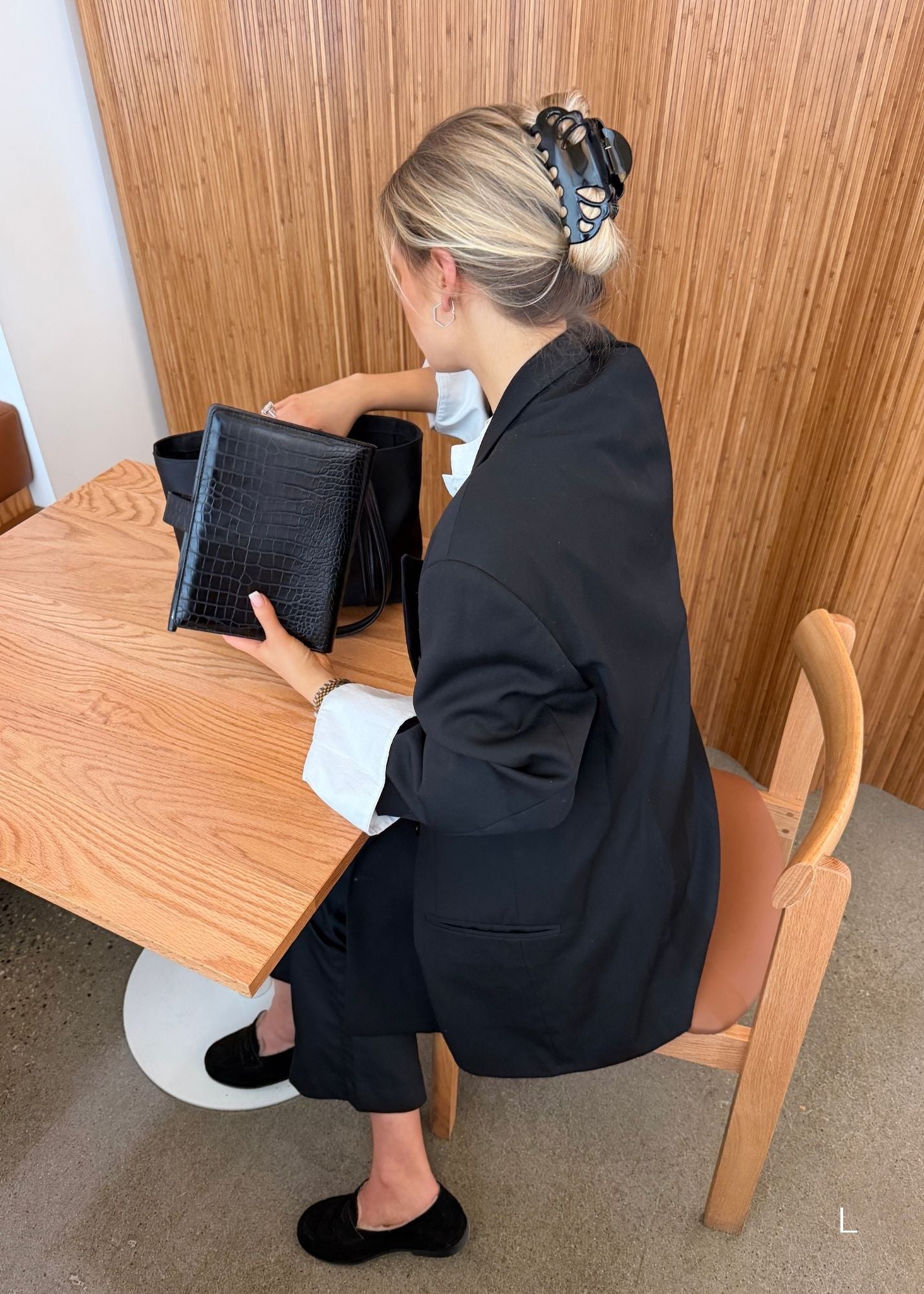 Woman sitting at a table with a black pouch, wearing a black outfit, with hair styled in an updo secured with a large black claw clip