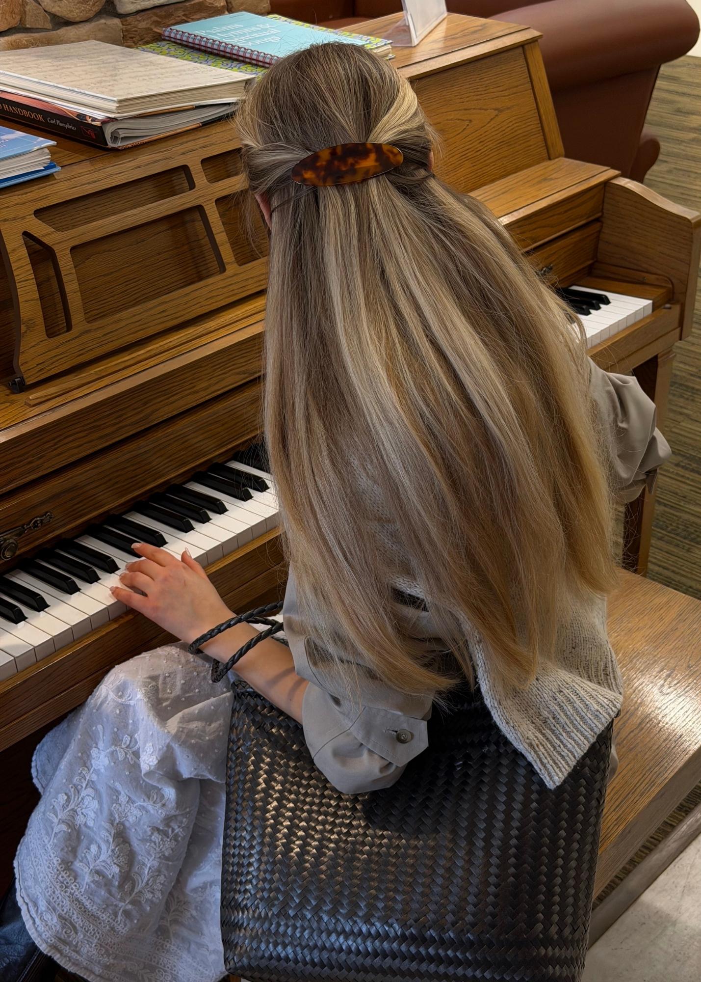 Woman playing a piano with long, blonde hair styled with a tortoise-coloured barrette, wearing a black woven bag