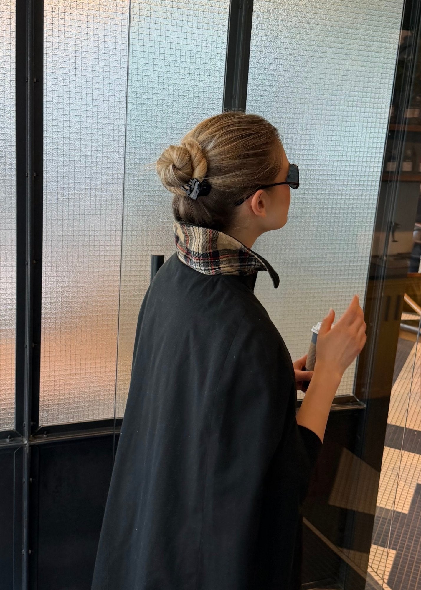 Woman wearing a black coat with a plaid collar, standing near a glass wall, with their hair styled in a bun secured by a small black claw clip