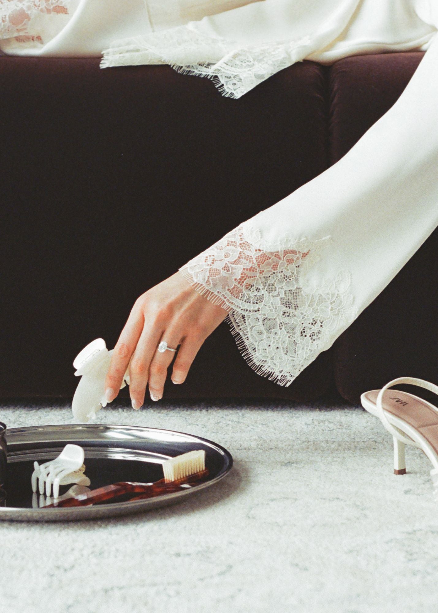 Woman laying on a brown couch in lace dress with white high-heeled shoes on a rug and a silver tray with 2 pearl coloured hair clips and a tortoise coloured fly away brush