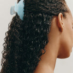 Woman with dark curly hair styled half-up, half-down with a small light blue claw clip against a neutral background