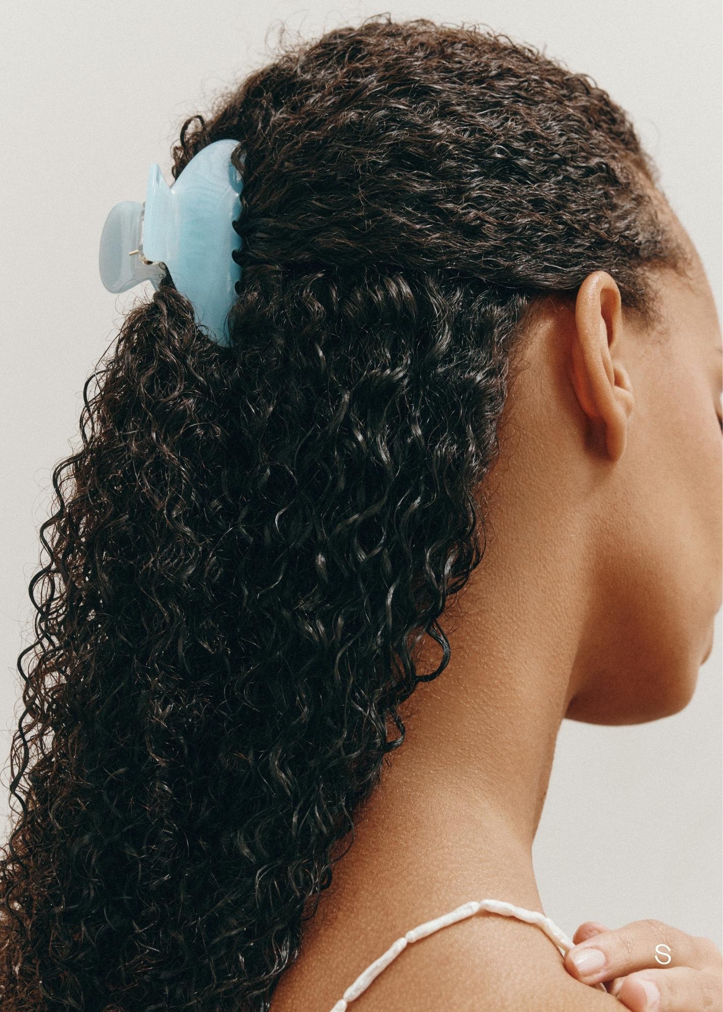 Woman with dark curly hair styled half-up, half-down with a small light blue claw clip against a neutral background