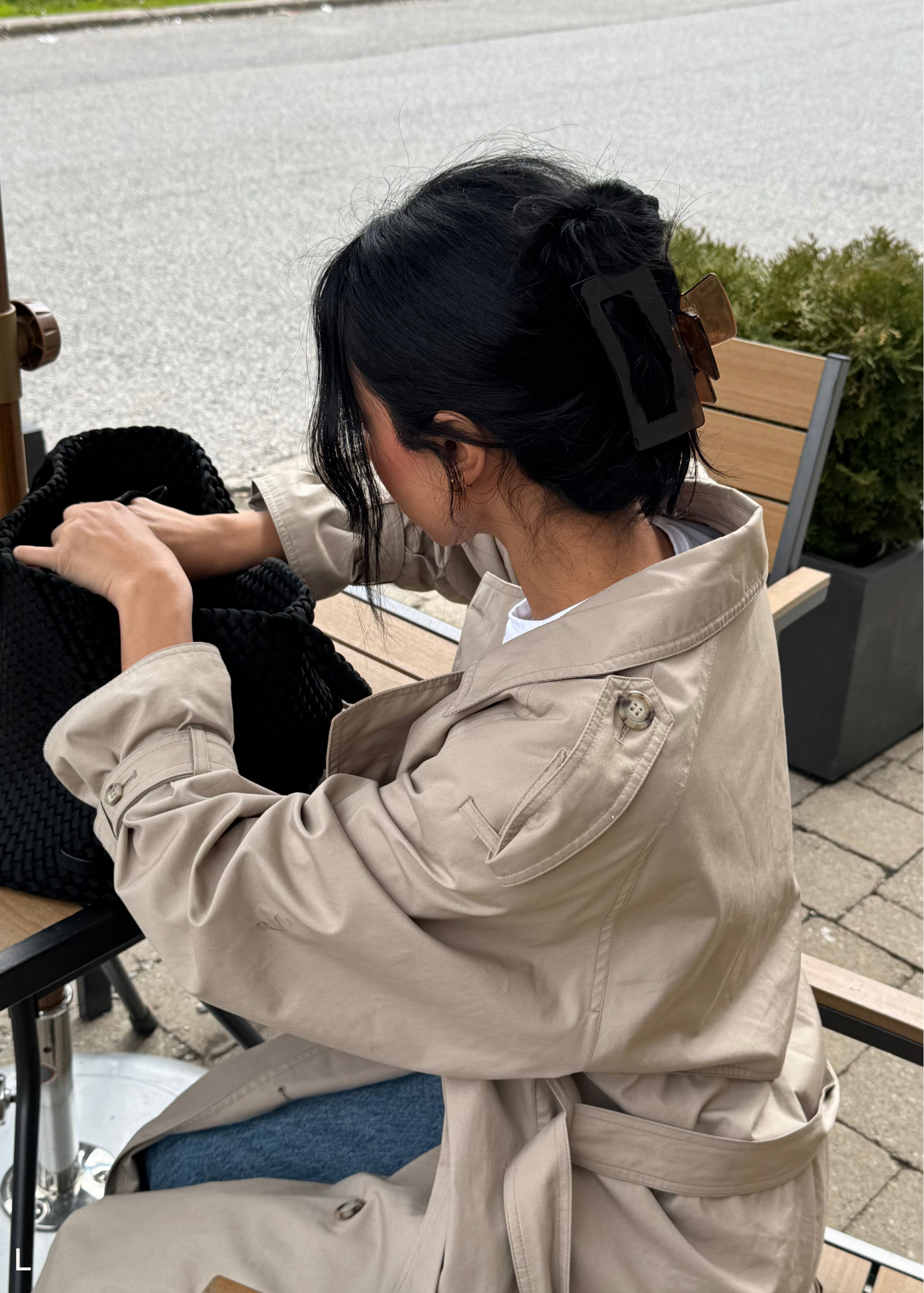 Person sitting outdoors, wearing a beige trench coat and a brown hair clip