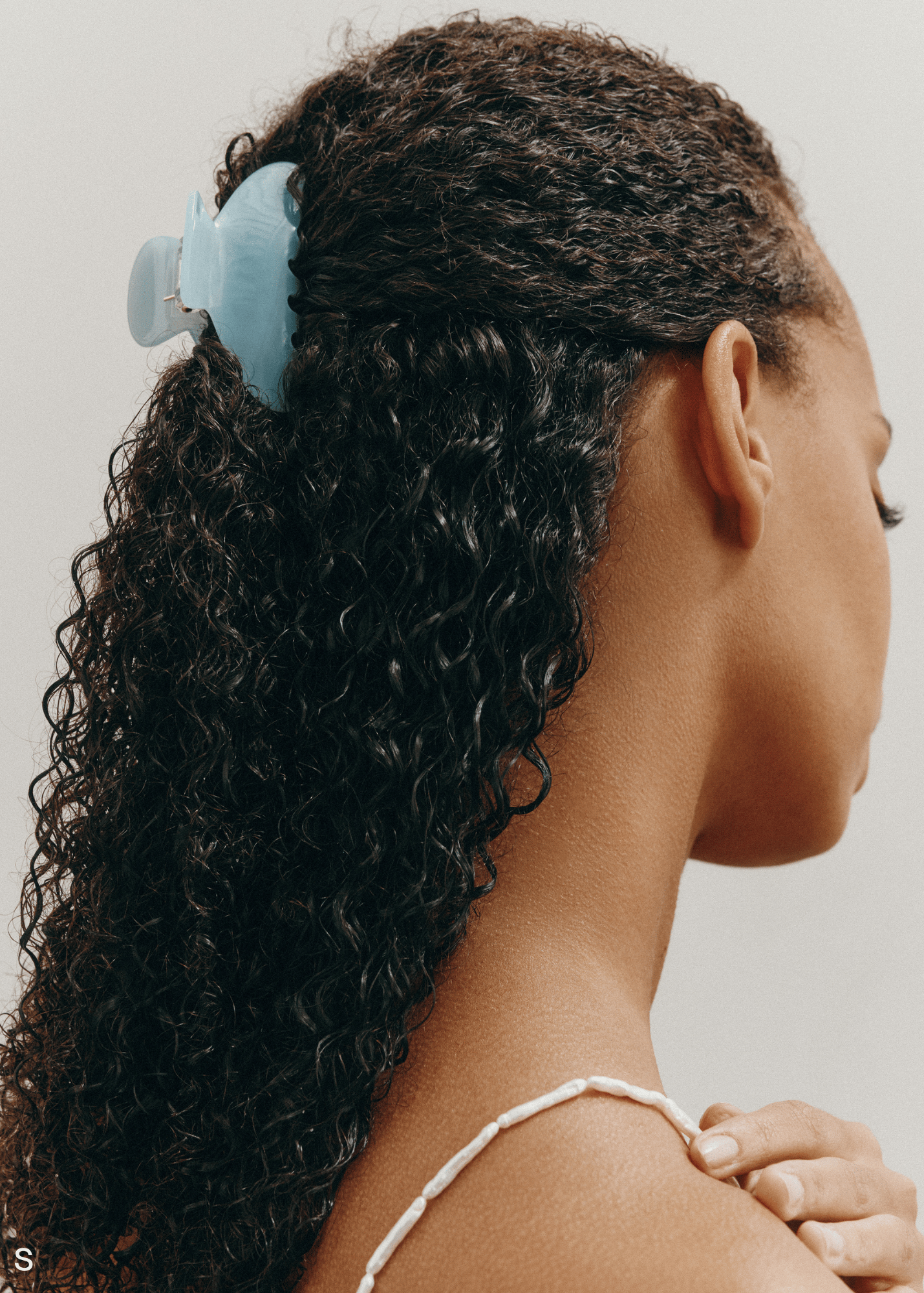 Woman with curly hair worn half up in a small light blue hair clip on a neutral background.