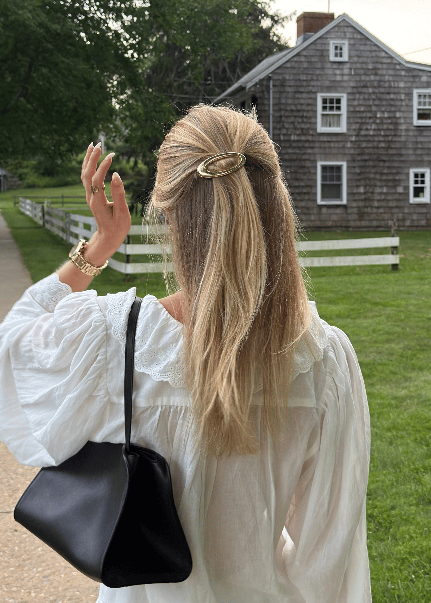 A woman with blonde hair tied back with a gold oval hair clip, wearing a white blouse and carrying a black shoulder bag, photographed from behind in an outdoor setting with a house and green lawn in the background.