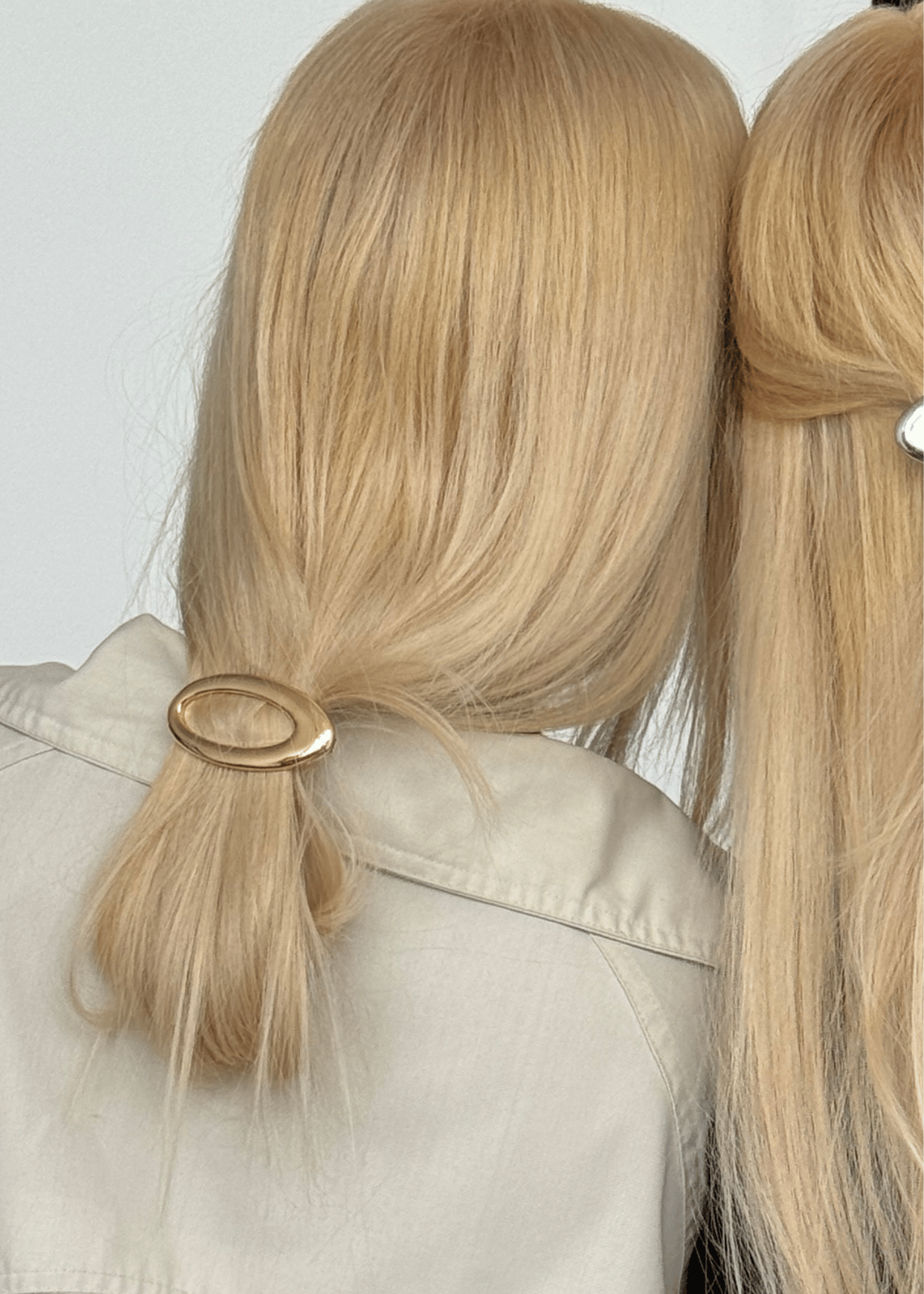 A close-up back view of a person with long blonde hair tied in a low ponytail with a gold oval hair accessory, wearing a light beige jacket against a plain light background.