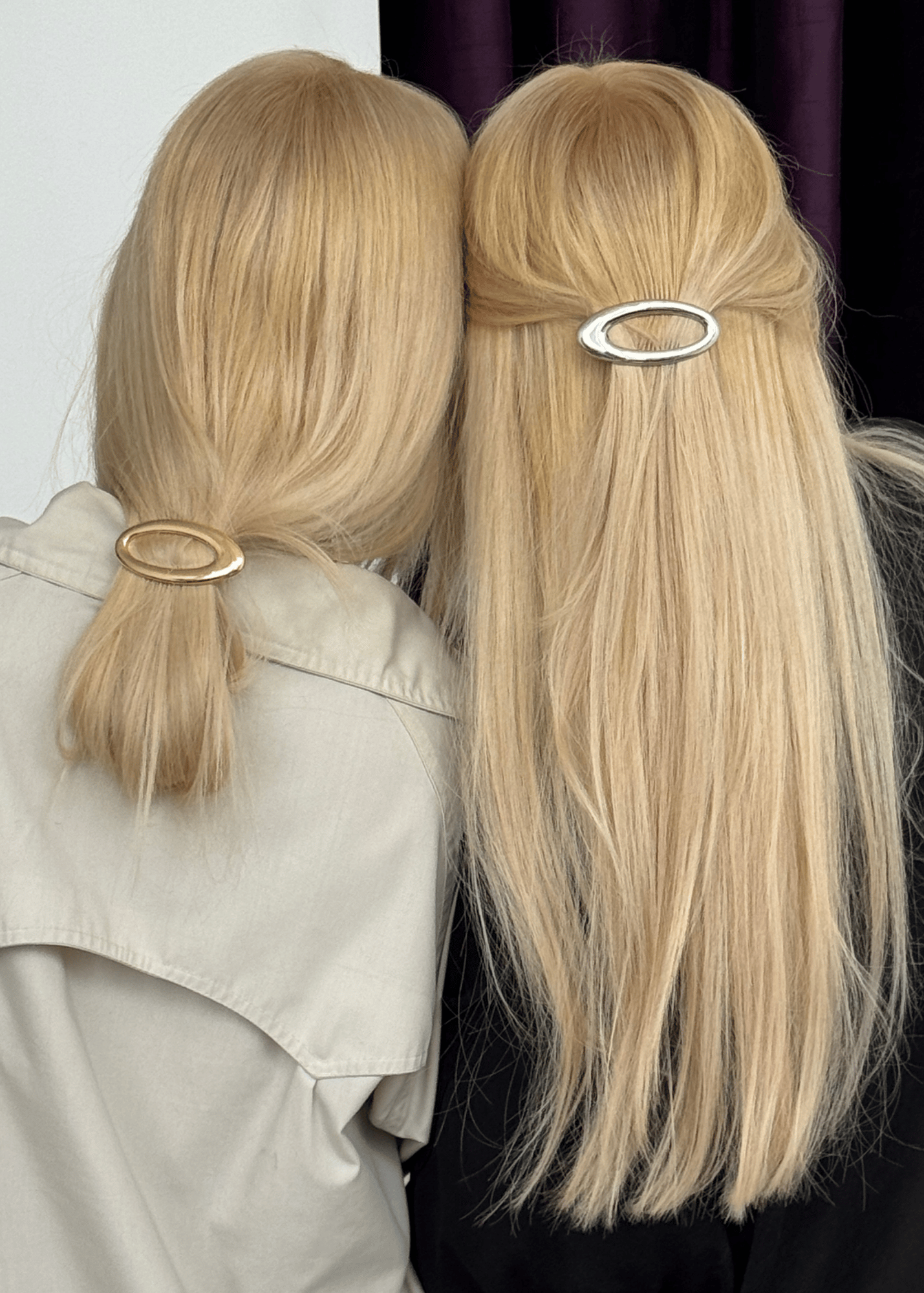 Two blonde women shown from behind wearing minimalist oval hair clips. The woman on the left wears a gold-toned clip holding her hair in a low ponytail, while the woman on the right wears a silver-toned clip holding her hair in a half-up style.