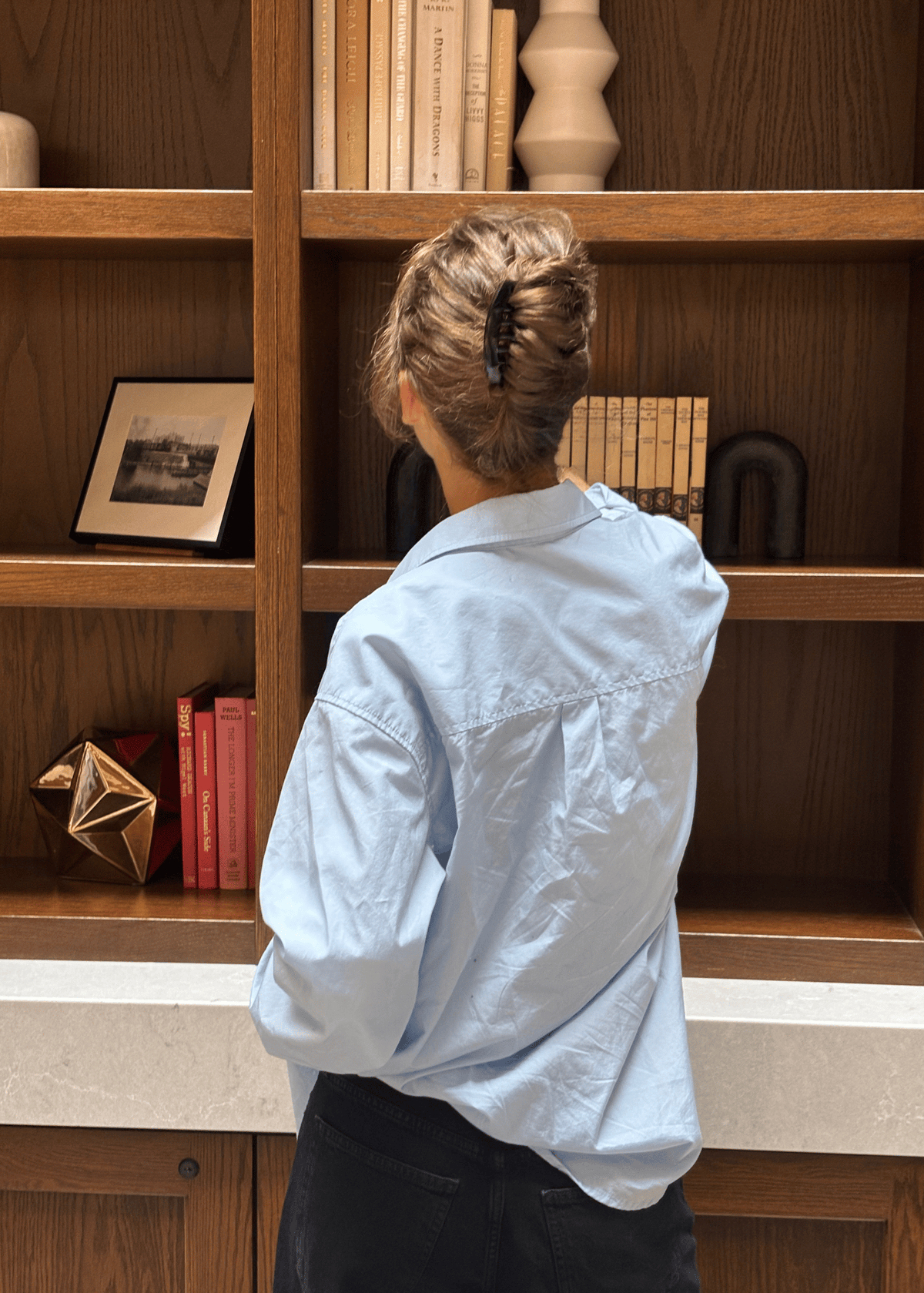 A woman shown from the back pulls a book off of a shelf. She is wearing her hair in a French Twist, secured by a black French comb. 