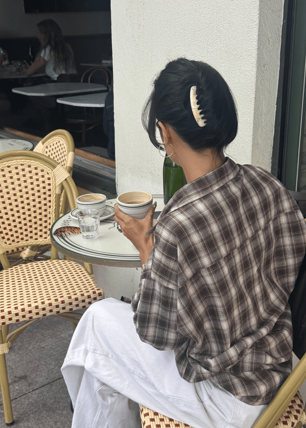 A woman sits at a cafe drinking coffee. Her hair is secured in a French twist by a light-neutral hair comb. 