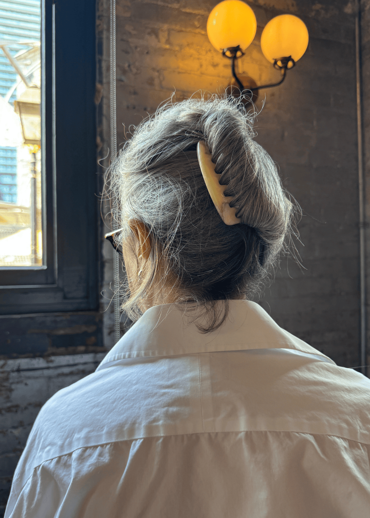 A grey-haired woman from the back wears a French twist hairstyle. It is securing using a light-neutral hair comb. 