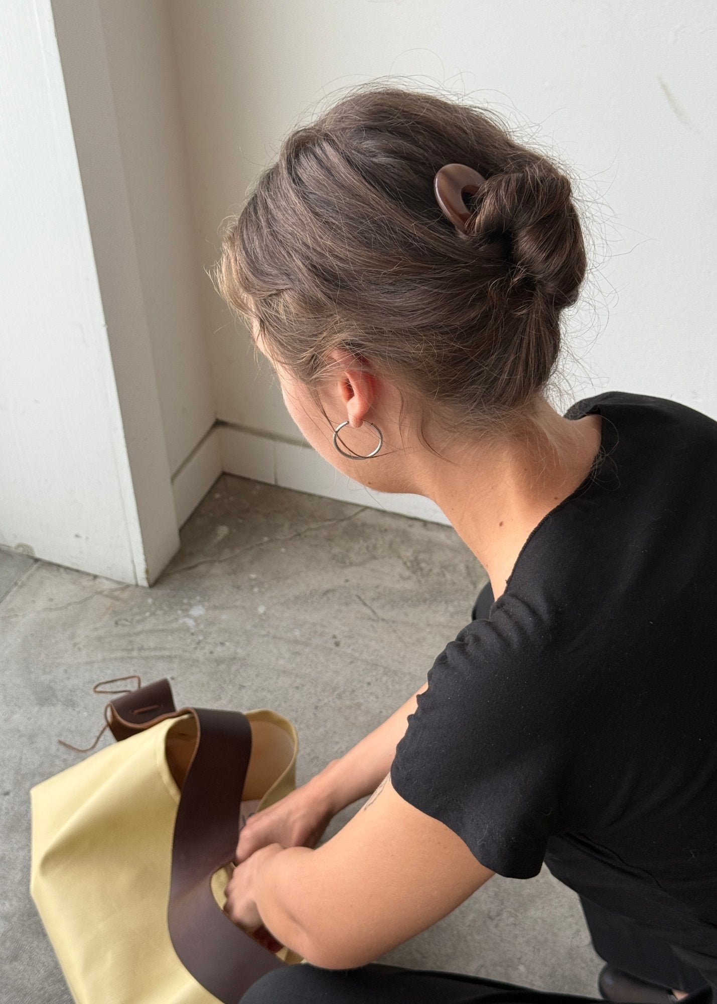 Woman sitting on the floor with a beige bag, wearing a black shirt, hoop earrings and a hair bun styled with a honey-coloured french pin