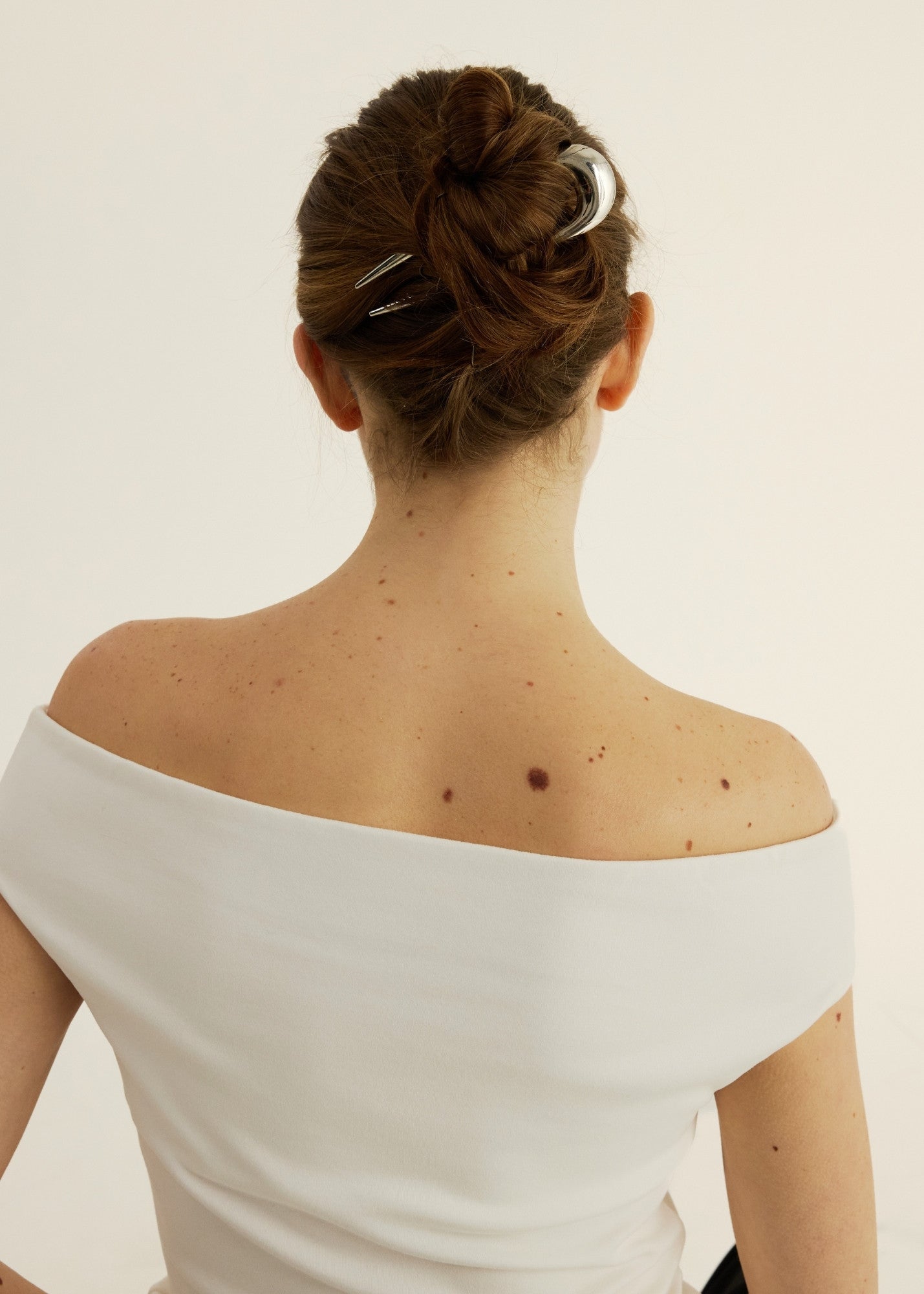Back view of a woman with a stylish hair bun secured with a silver french pin wearing a white off-shoulder top with a neutral background