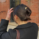 Woman with a bun secured with the French Pin in Gold and earrings adjusting their hair against a brick building with gold numbers.
