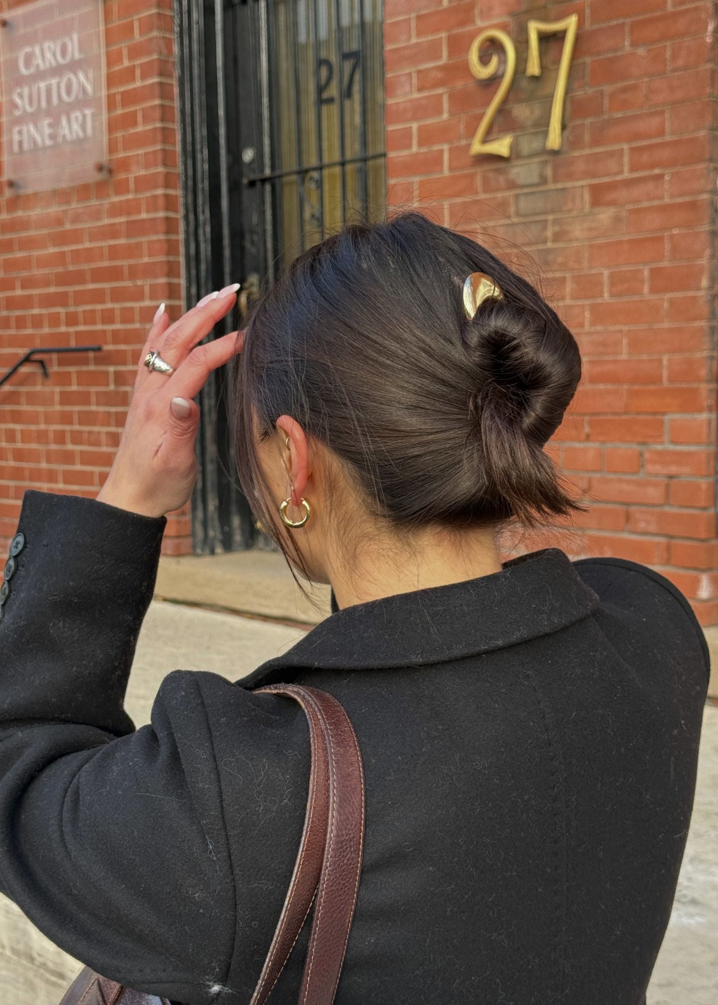 Woman with a bun secured with the French Pin in Gold and earrings adjusting their hair against a brick building with gold numbers.