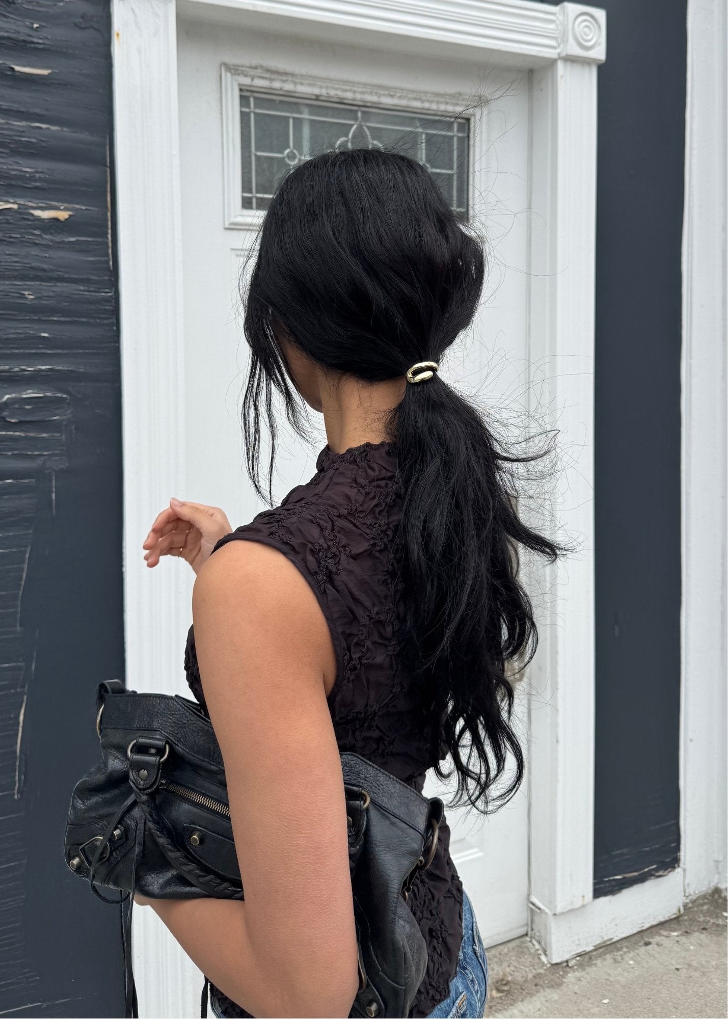 Person with long black hair tied back styled with a small, gold hair accessory, wearing a dark top and carrying a black bag, standing in front of a white door.