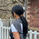 Woman with long black hair in a ponytail secured with a honey-coloured barrette sitting outdoors near a white fence and brick building.