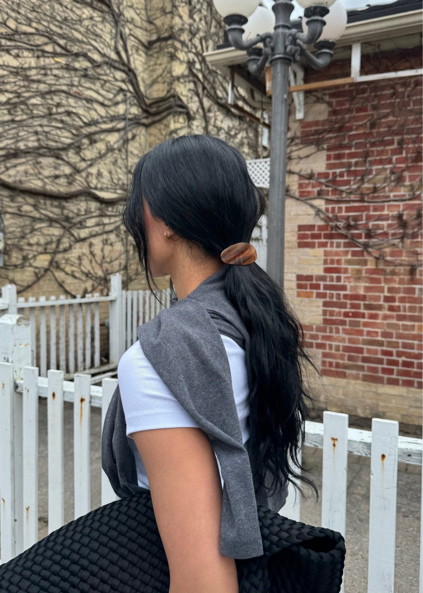 Woman with long black hair in a ponytail secured with a honey-coloured barrette sitting outdoors near a white fence and brick building.