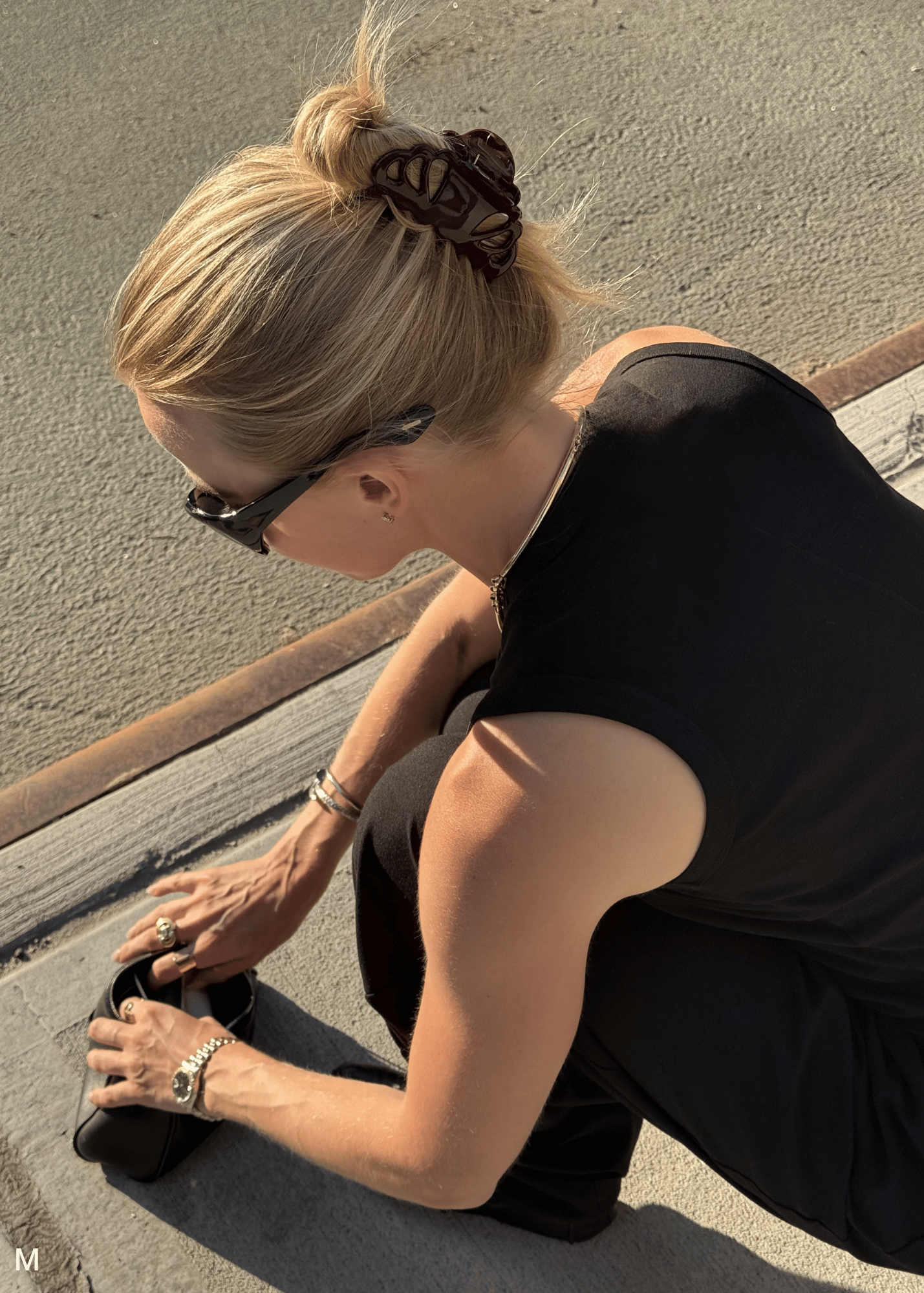 A woman in a black sleeveless top and black pants crouches outdoors on a concrete surface, looking though her bag. She has blonde hair in a bun with a deep purple hair clip, wears black sunglasses, and has bracelets on her wrists.