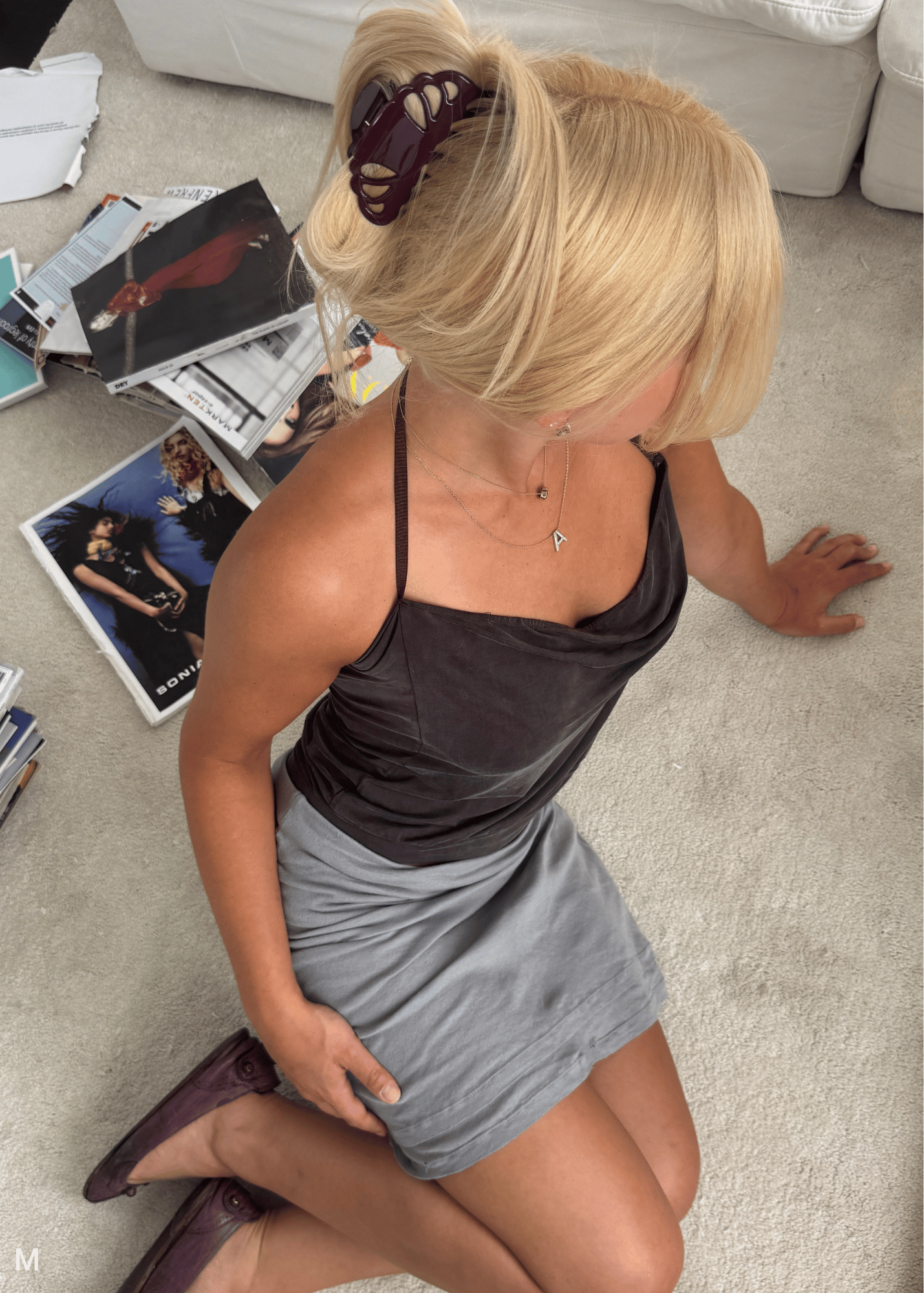A woman with blonde hair in a deep purple claw clip sits on a light beige carpet, wearing a dark brown satin camisole top and a light gray mini skirt. She is viewed from an overhead angle, with magazines scattered on the floor around her.