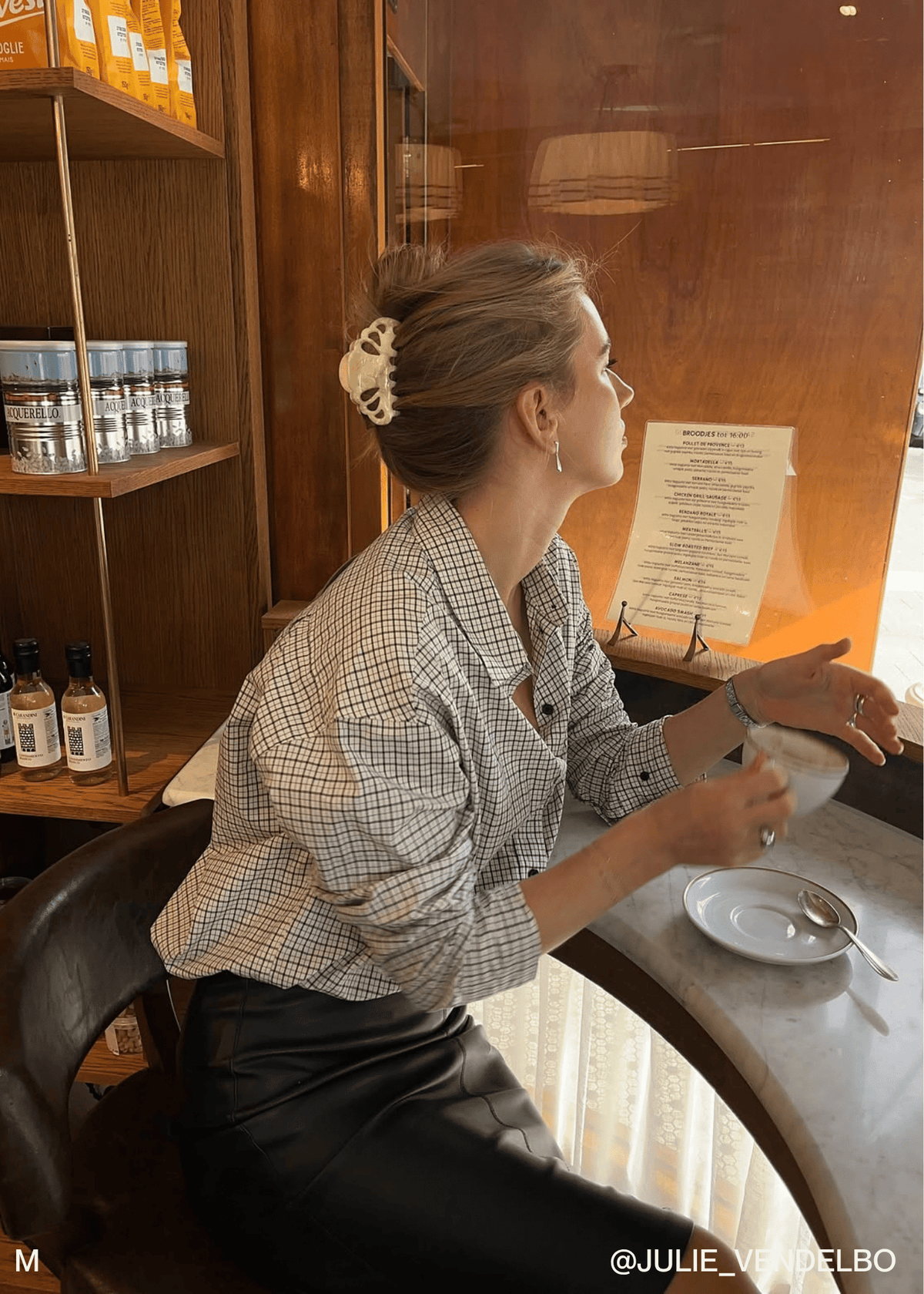 A woman sits at a cafe table wearing a light beige and black checkered button-up shirt and black leather pants, holding a white cup. She has her hair in a bun with a cream-colored claw clip.