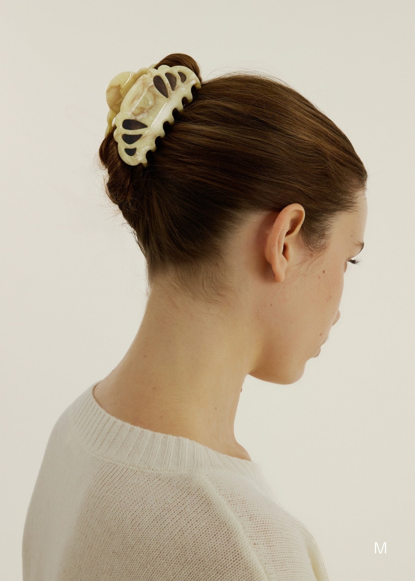 Person with long hair in a hairstyle with a small, beige coloured hair clip with scalloped edges and tear drop shaped cut outs against a plain background.