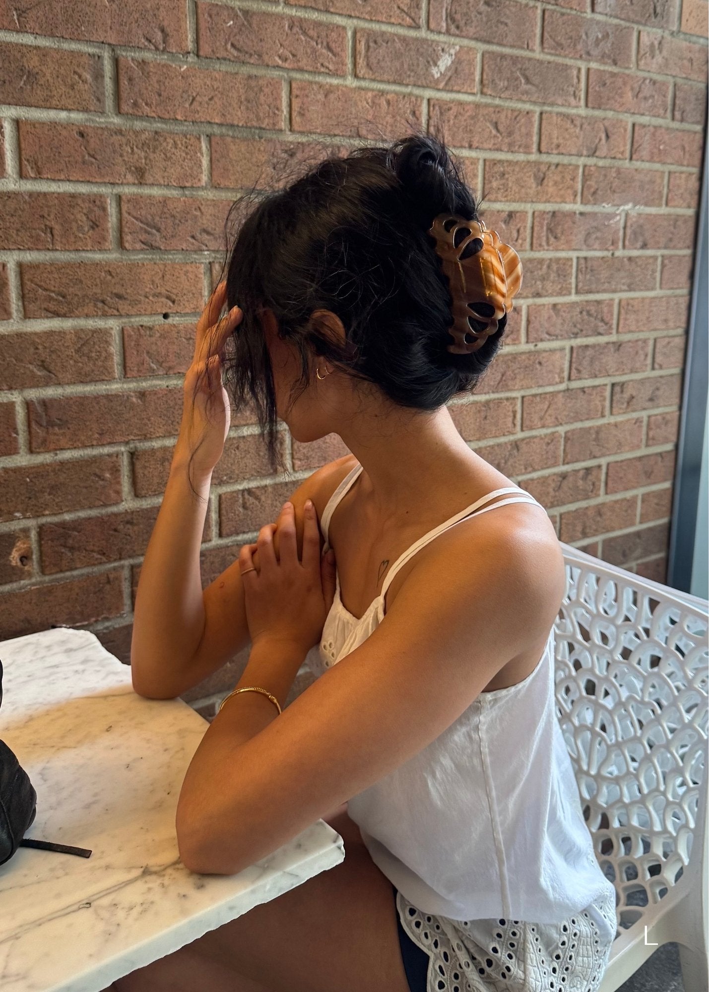 Woman with dark hair styled with a large honey-coloured decorative claw clip sitting on a bench with a brick wall background