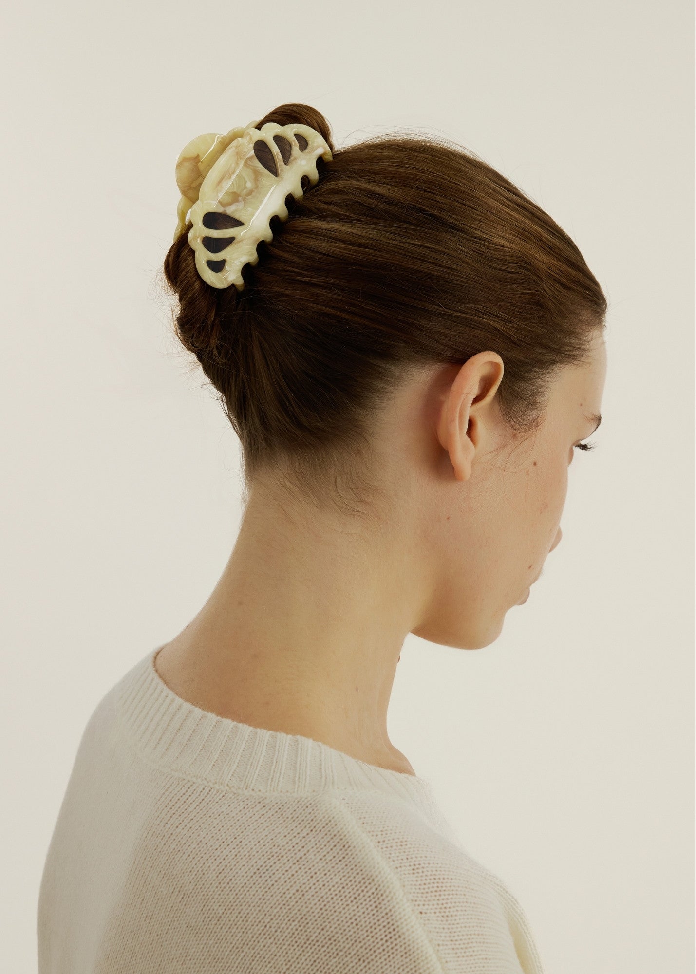 Person with long hair in a hairstyle with a small, beige coloured hair clip with scalloped edges and tear drop shaped cut outs against a plain background.