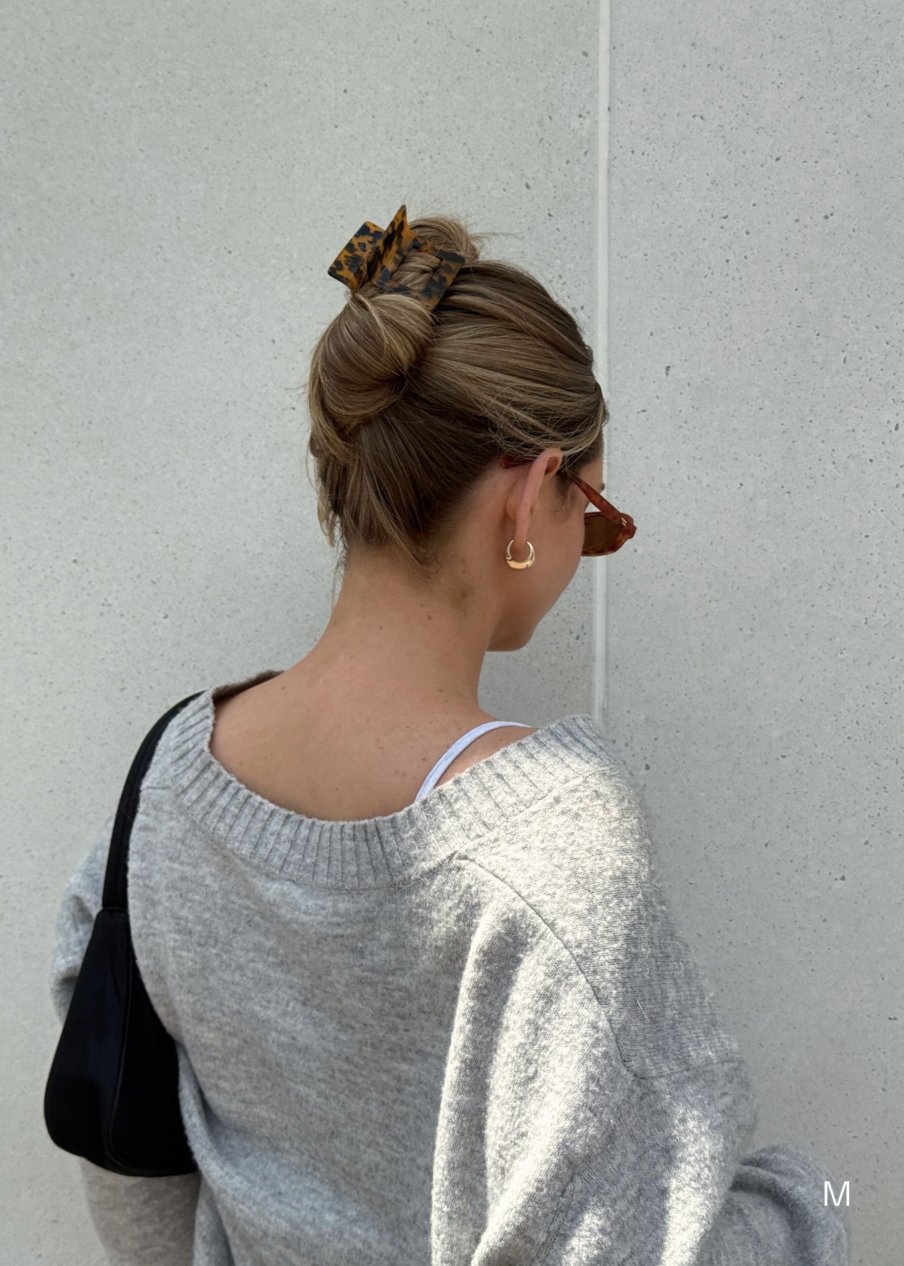 Woman in a gray sweater standing in front of  a light gray wall with an up-do hairstyle secured with a dark tortoise coloured hair clip