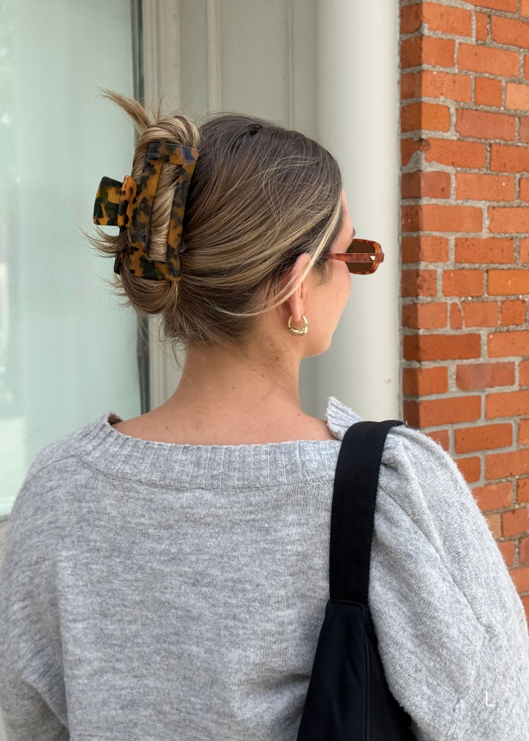 Person with a large dark tortoise coloured hair clip in their hair and wearing a grey sweater, standing against a brick wall.
