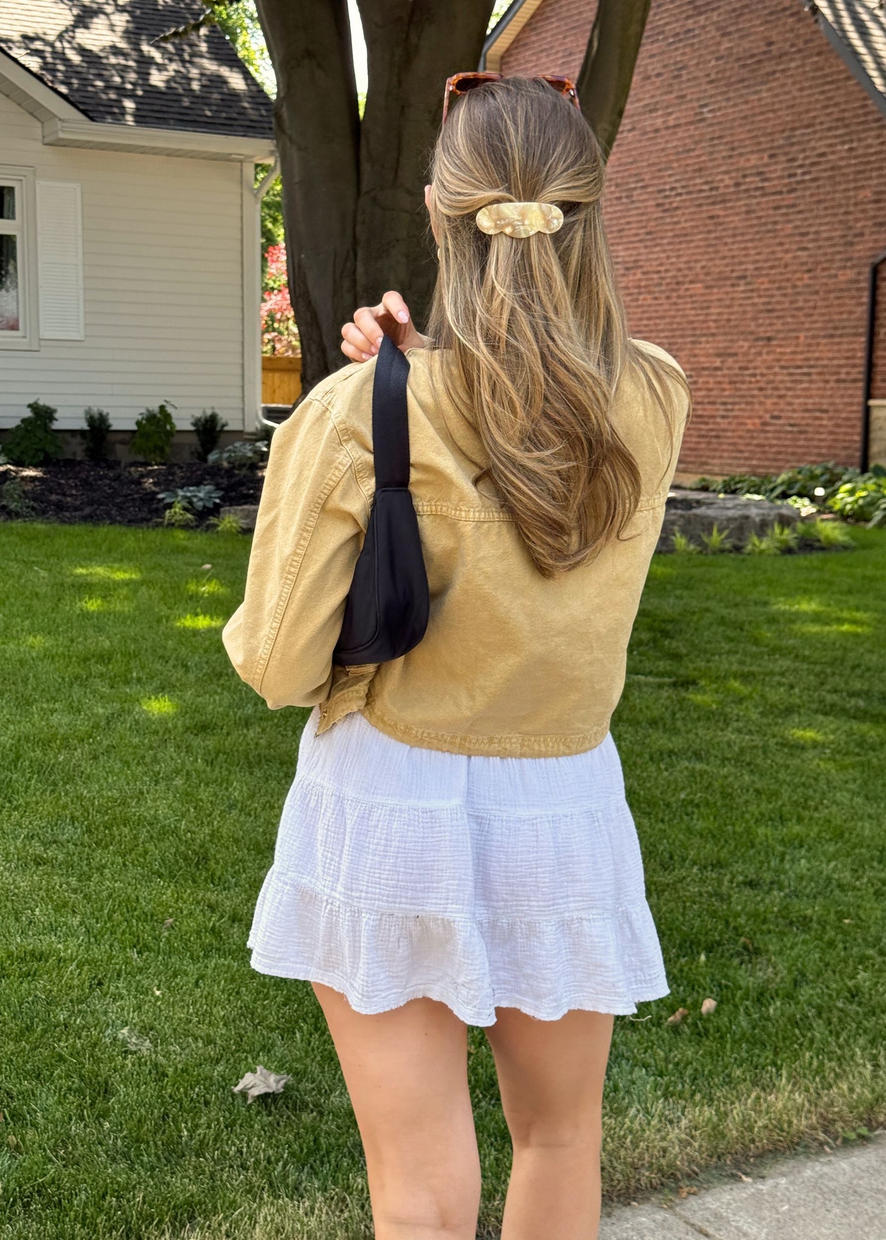 Person wearing a jacket and white skirt standing in a grassy area with houses in the background showing their half-up, half-down hairstyle secured with a scalloped edge barrette in a beige colour.
