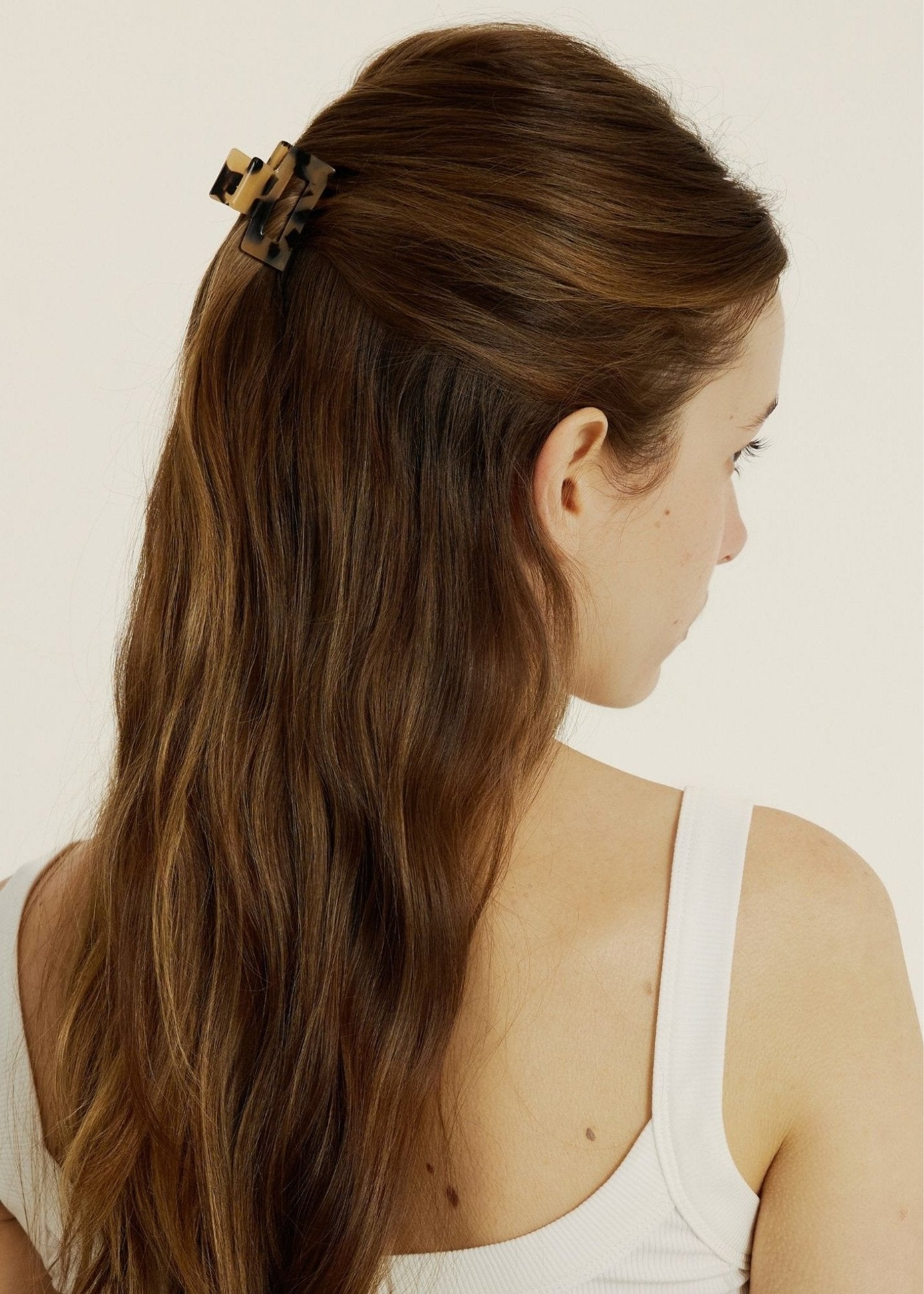 Woman with long brown hair styled half-up, half-down secured with a mini hair clip wearing a white tank top against a plain background