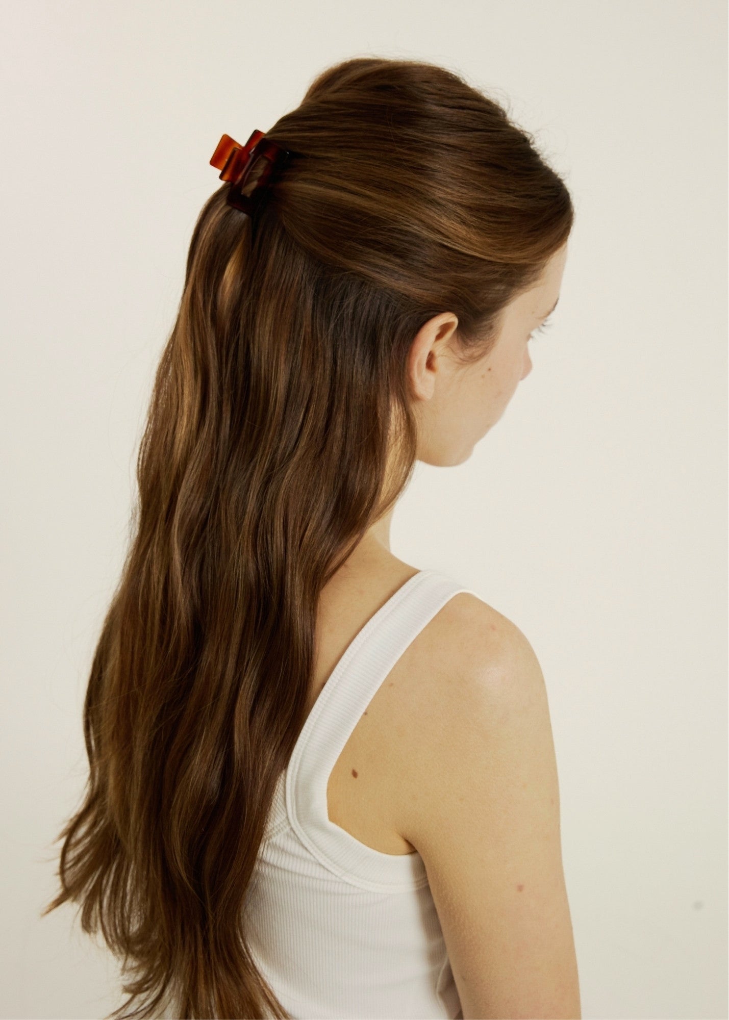 Woman with long brown hair styled half-up, half-down with a mini reddish coloured hair clip wearing a white tank top against a plain background.