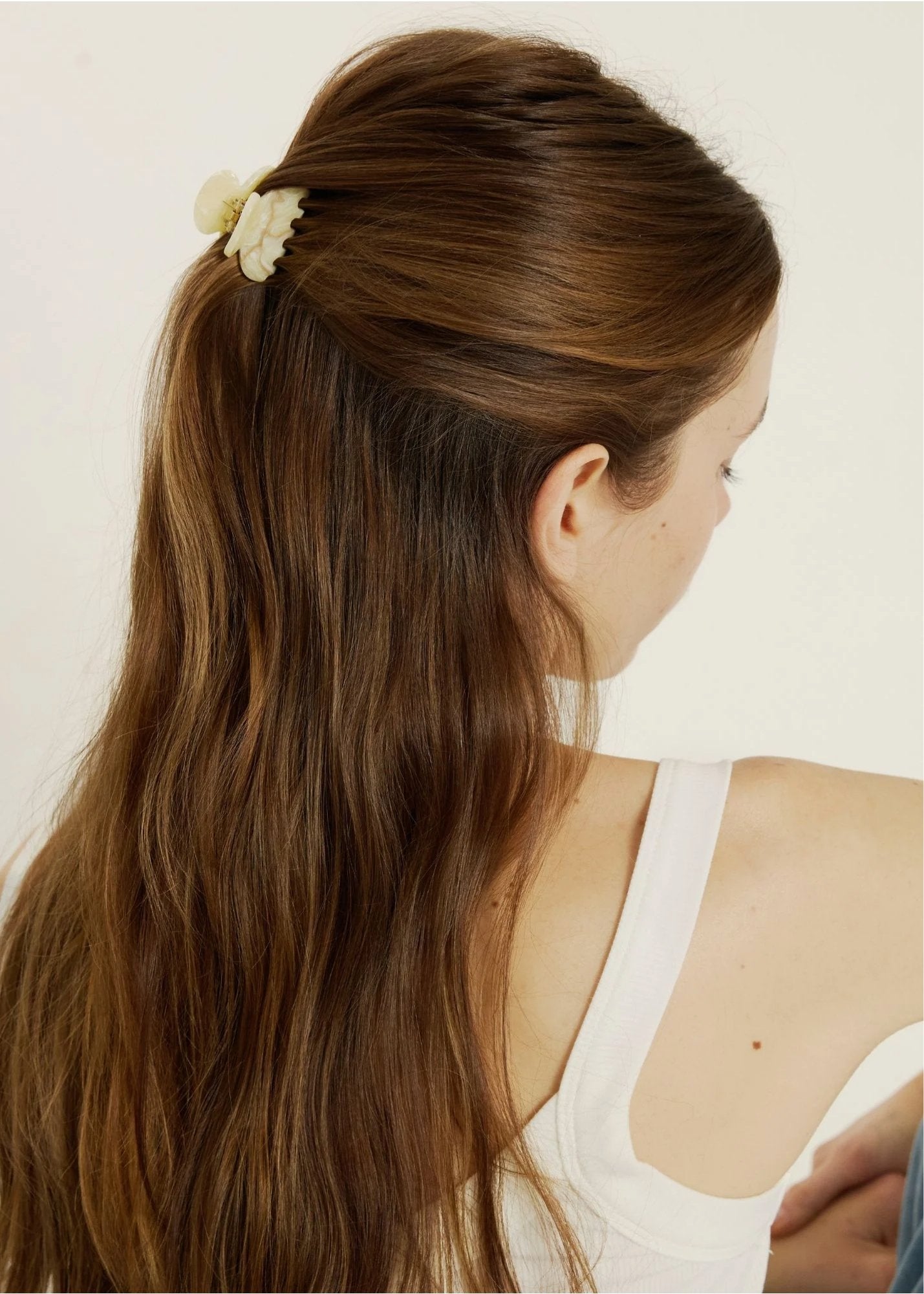 Woman with long brown hair styled in a ponytail with a minil beige-coloured hair clip, wearing a white tank top.