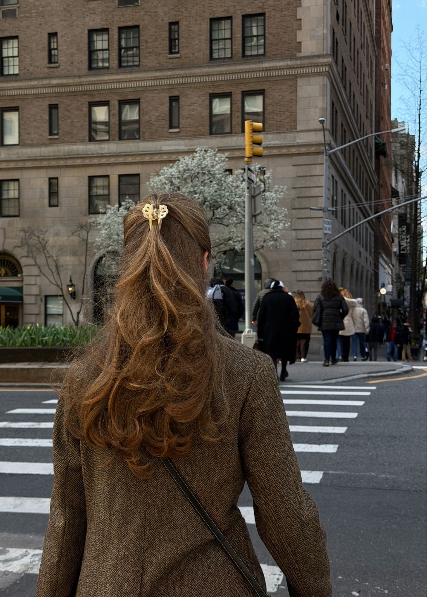 Woman with long brown hair wearing a small beige-coloured hair clip crossing a city street.