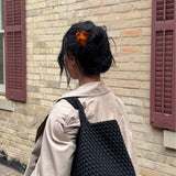 Woman with dark hair styled in a bun secured by a decorative tortoise-coloured claw clip wearing a woven black bag walking past a brick wall with purple shutters.