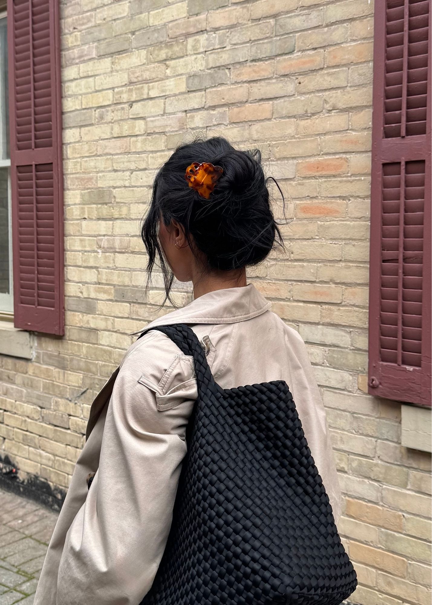 Woman with dark hair styled in a bun secured by a decorative tortoise-coloured claw clip wearing a woven black bag walking past a brick wall with purple shutters.