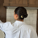 Back view of woman with dark hair styled in a bun styled by a beige-coloured french pin wearing a white dress shirt in a kitchen setting with wooden cabinets and tiled wall.