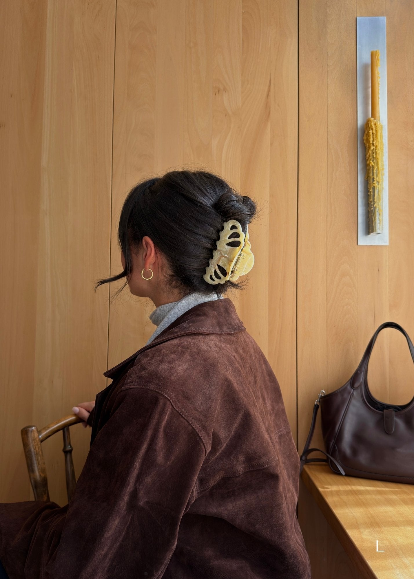 Woman with dark hair styled with a beige coloured claw clip, sitting in a room with wooden walls and a handbag on a bench.