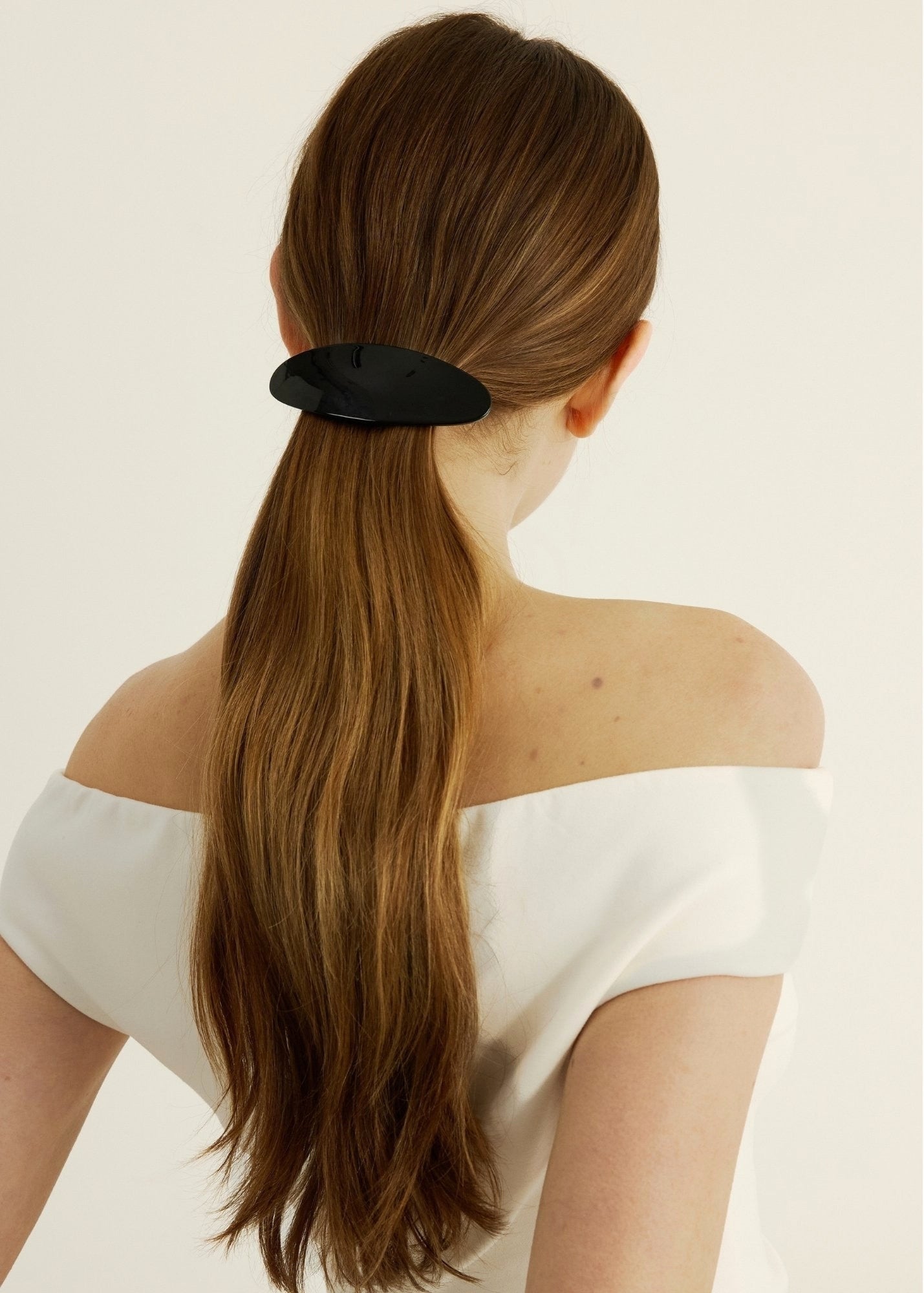 Woman with long brown hair secured back with a black barrette, wearing a white off-shoulder top.