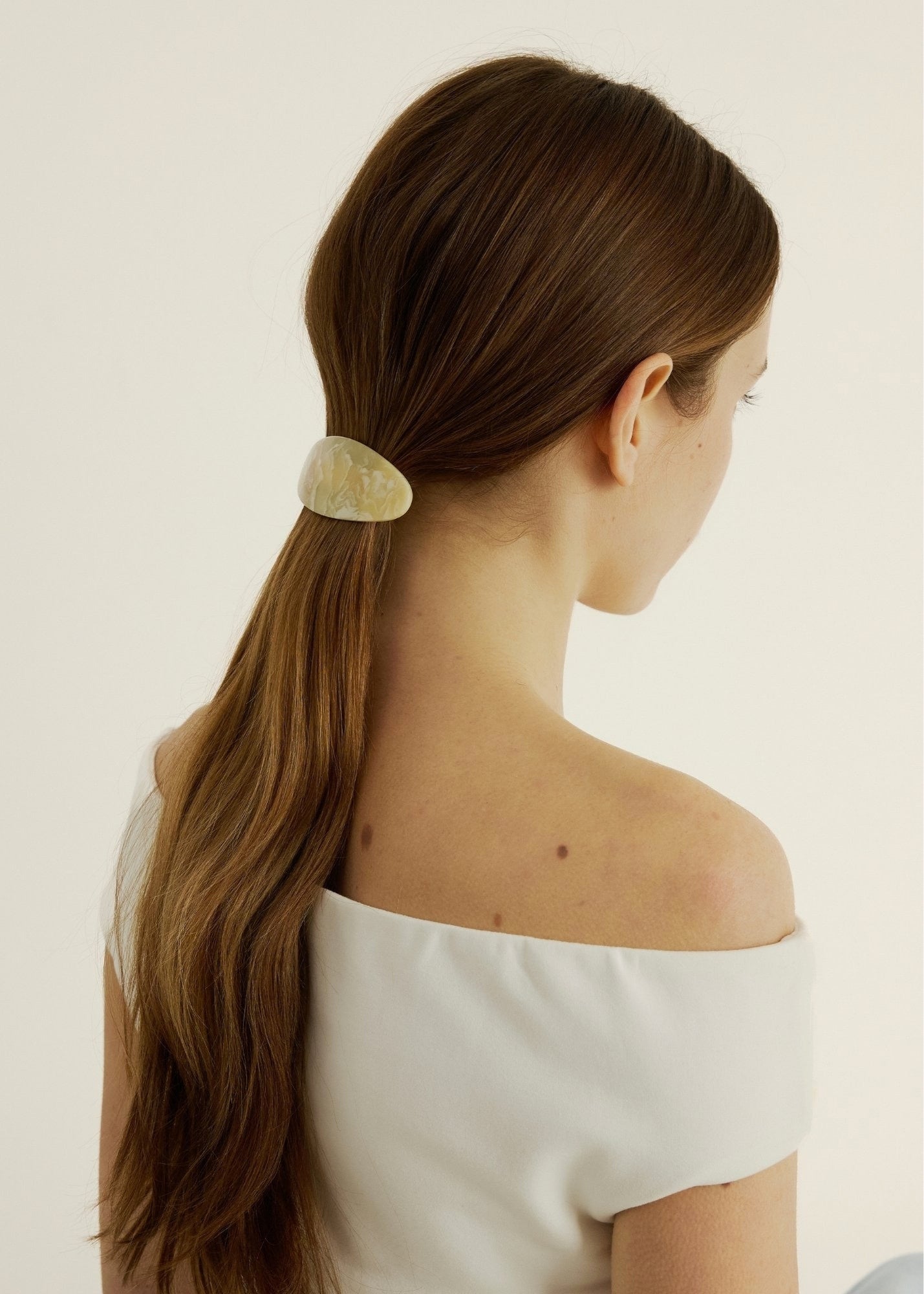 Woman with long brown hair secured back with a beige-coloured barrette against a plain background