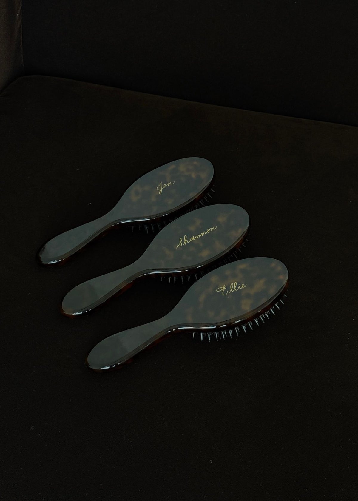 Three Refine hairbrushes in tortoise with names engraved in gold, cursive writing on a brown couch