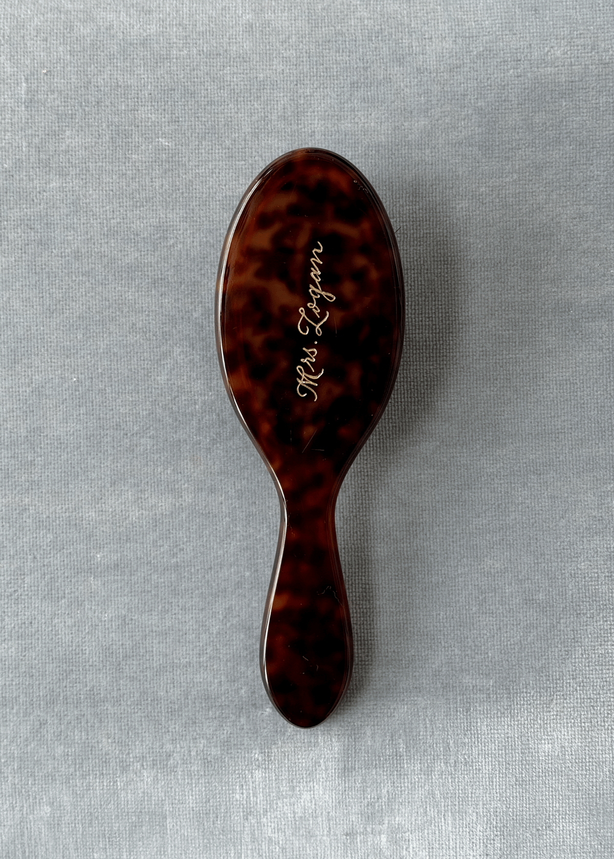 A hairbrush with a glossy dark brown tortoiseshell pattern and gold cursive script reading 'Mrs. Logan' on the back, set against a textured blue fabric background.