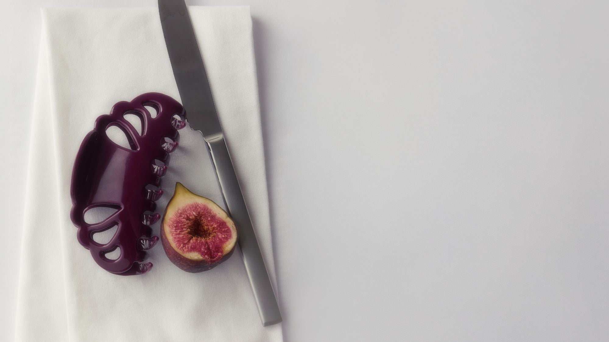A dark purple claw hair clip  displayed on a white napkin next to a silver knife and a halved fig on a light gray background.