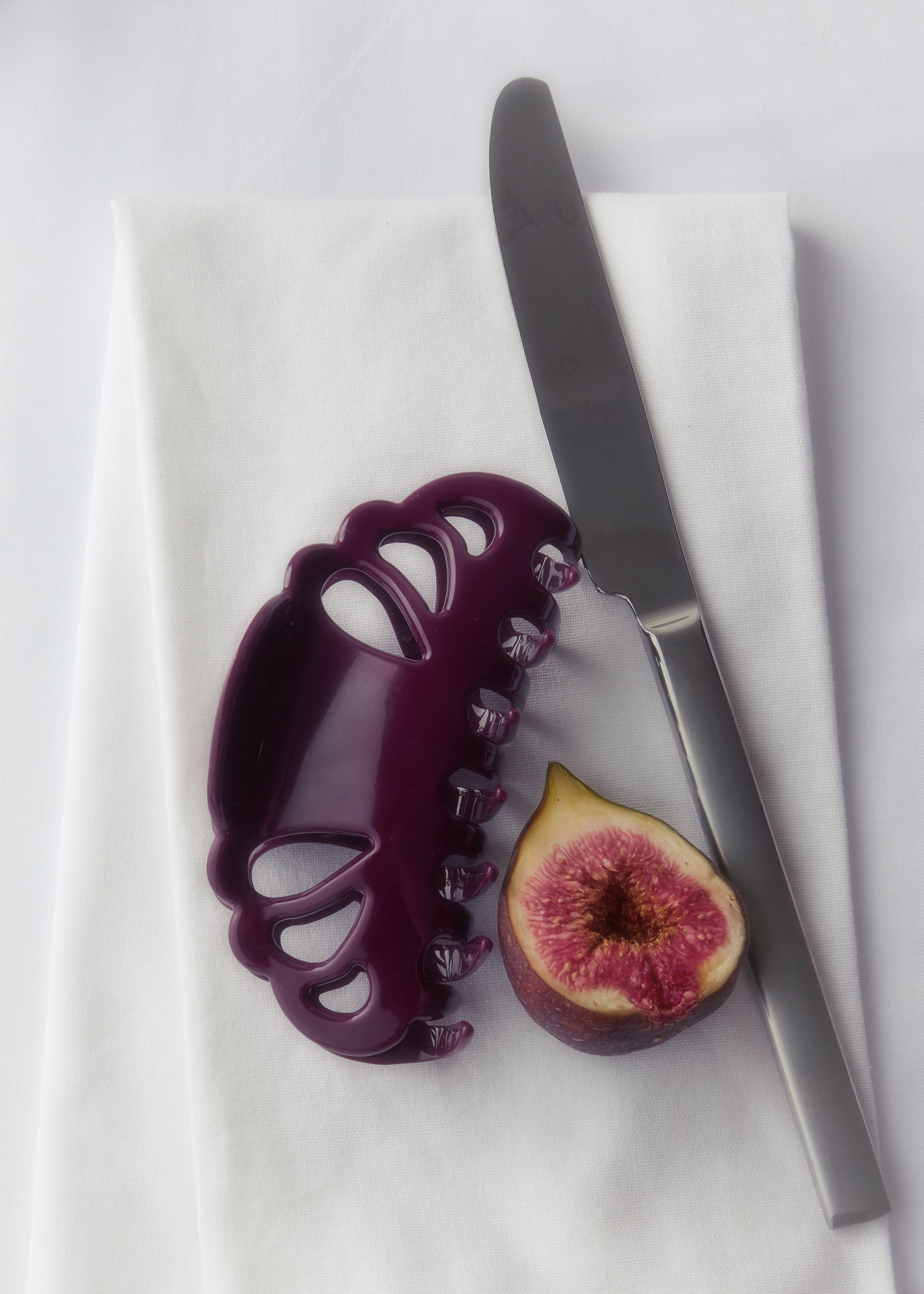 A dark purple plastic hair claw clip with decorative cutouts, displayed on a white cloth next to a silver knife and a halved fig.