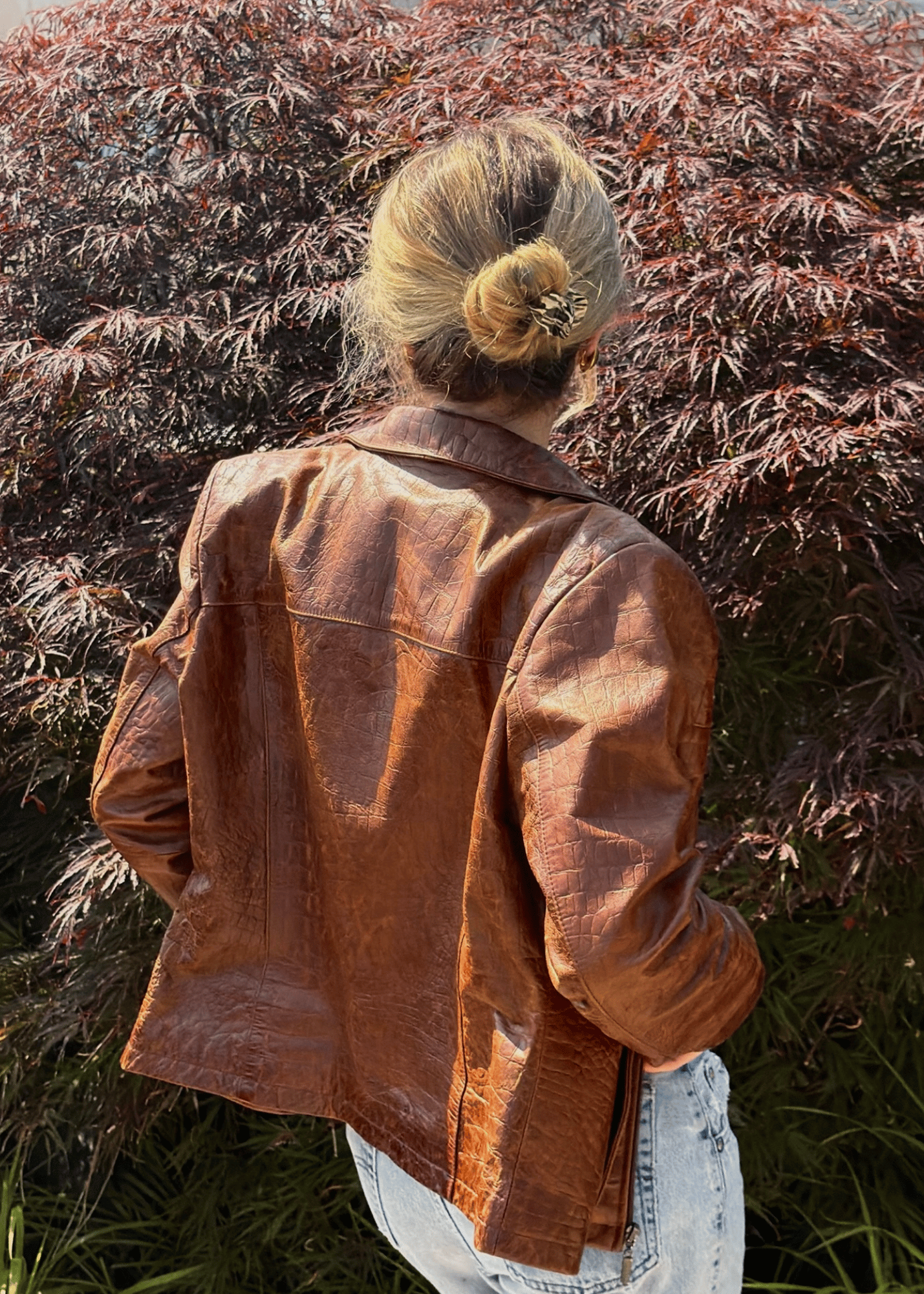 A woman viewed from behind wearing a brown textured faux-leather jacket and a small marbled claw clip, standing outdoors in front of reddish-purple foliage.