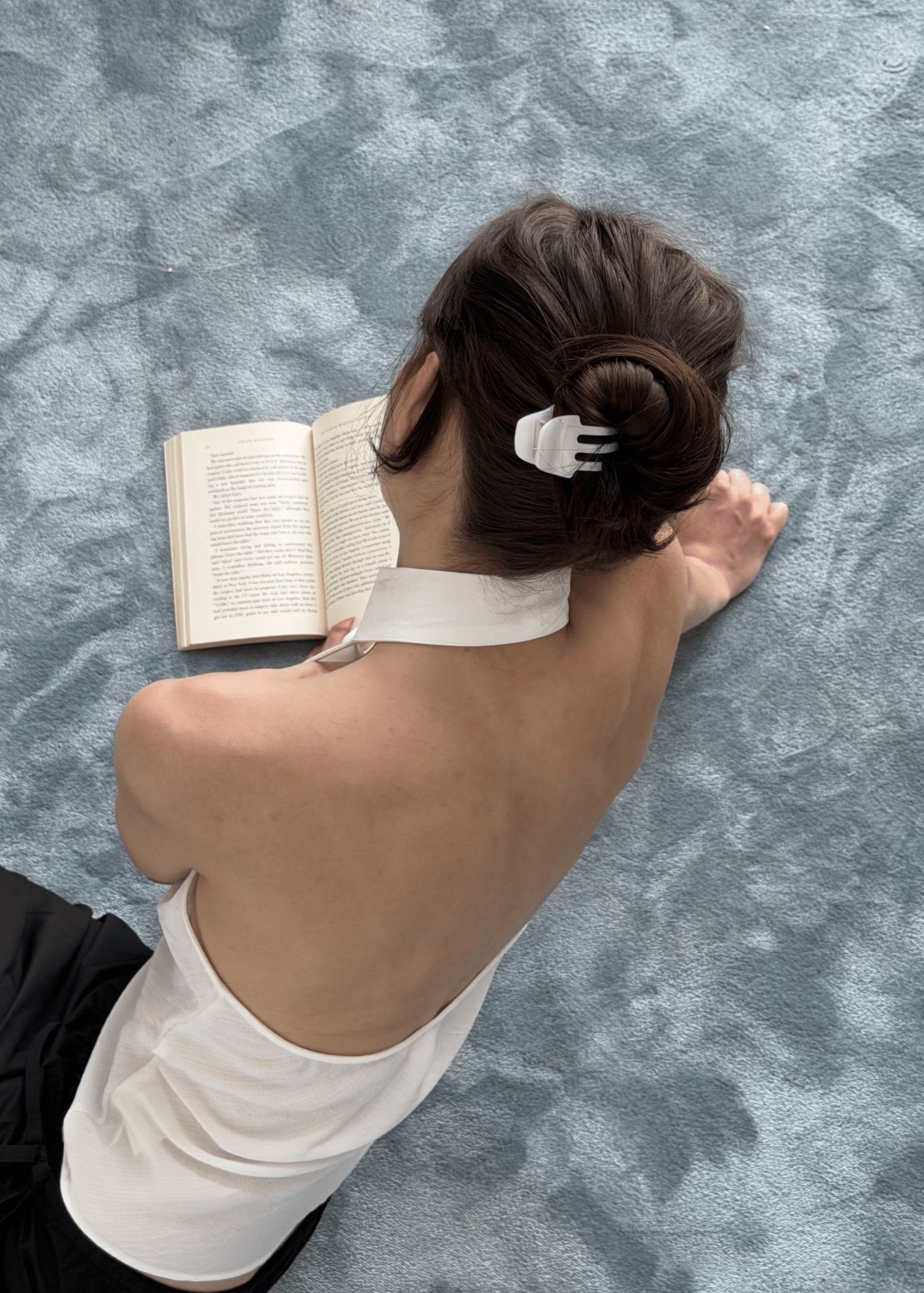 A woman viewed from behind sits on a textured blue-gray carpet, reading an open book. She has her hair in a bun secured with a white pearlescent claw clip and is wearing a white halter-style top with a high collar.